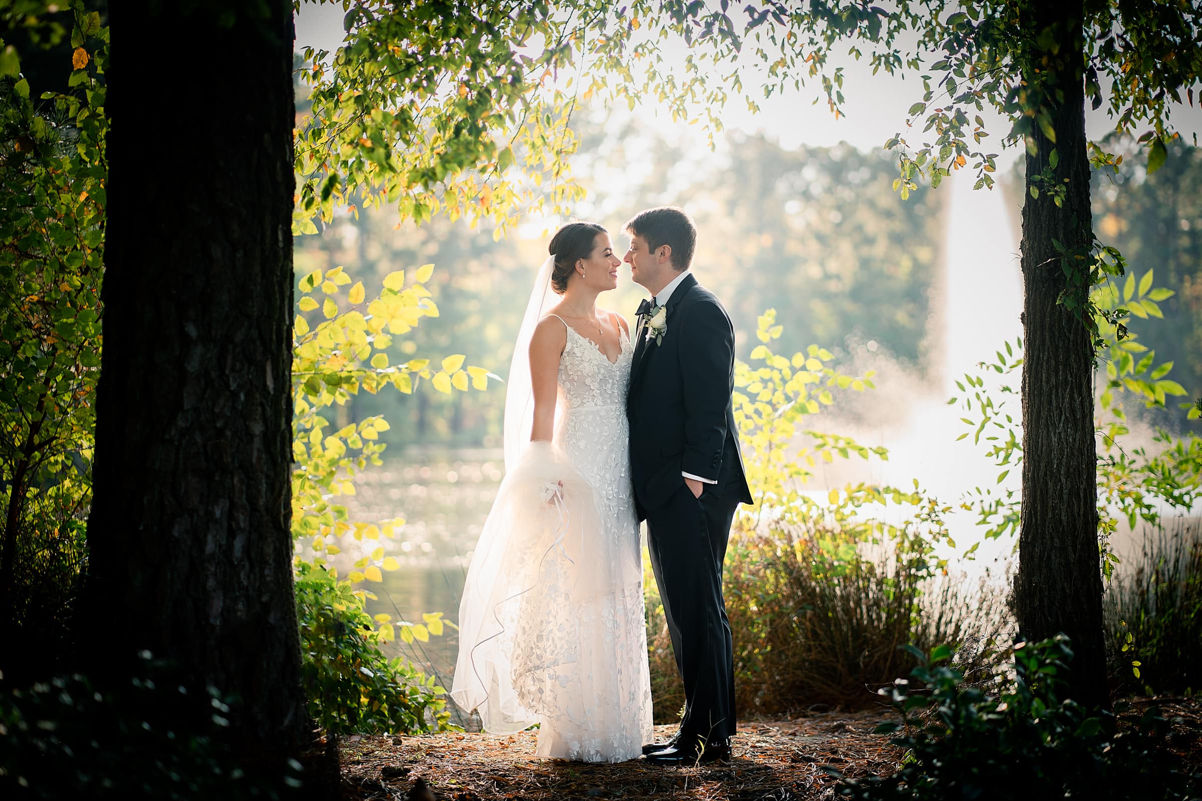 A bride and groom stand closely under trees, with the bride in a white dress and veil and the groom in a black suit and tie. The sun filters through the leaves, creating a romantic and serene backdrop near a pond.