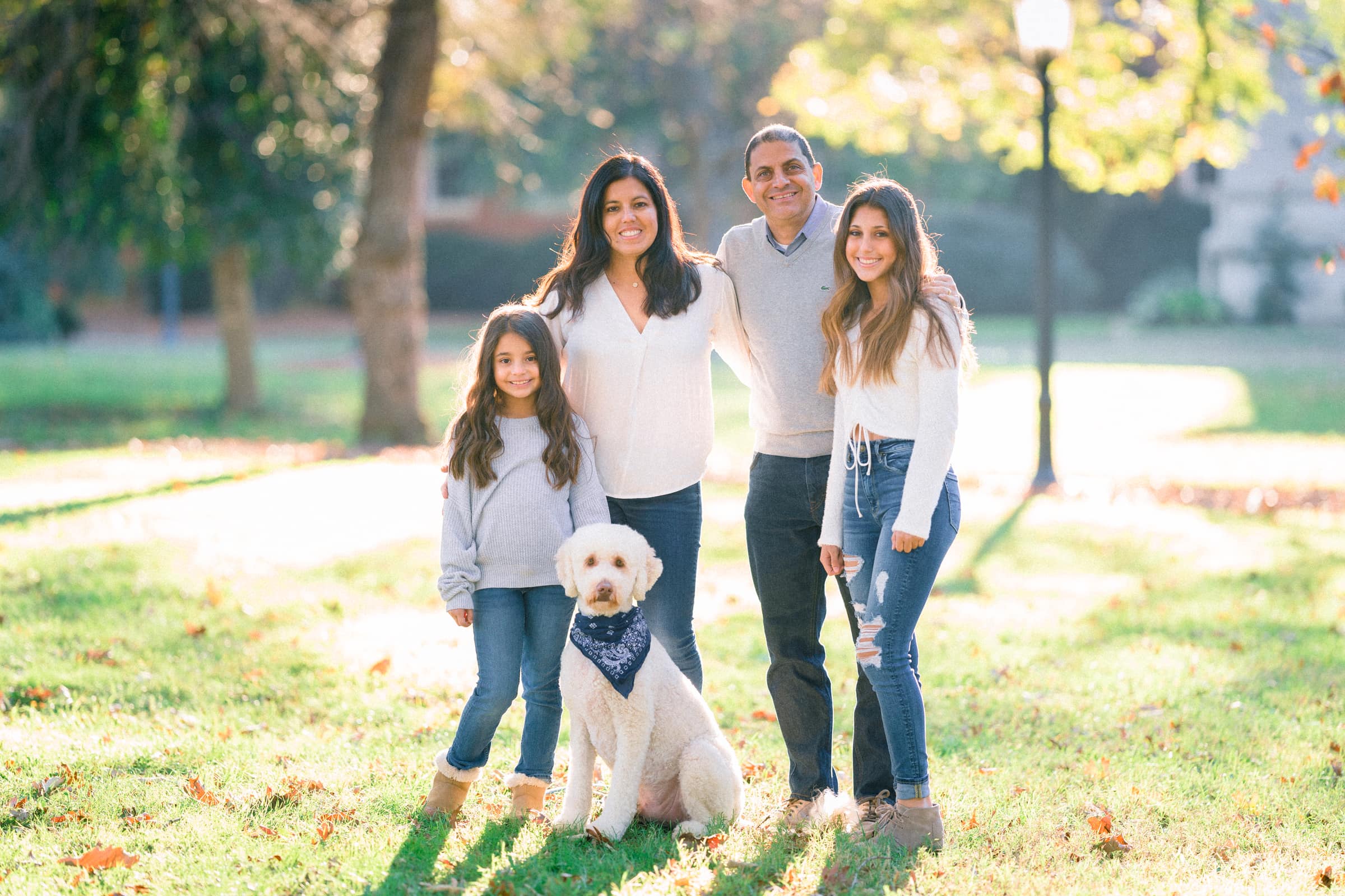 raleigh family photography - jackie, cesar & family A happy family of four, along with a white dog wearing a blue bandana, poses together in a sunlit park. The family, dressed in casual attire, smiles while standing on a grassy area with trees and a lamp post in the background.