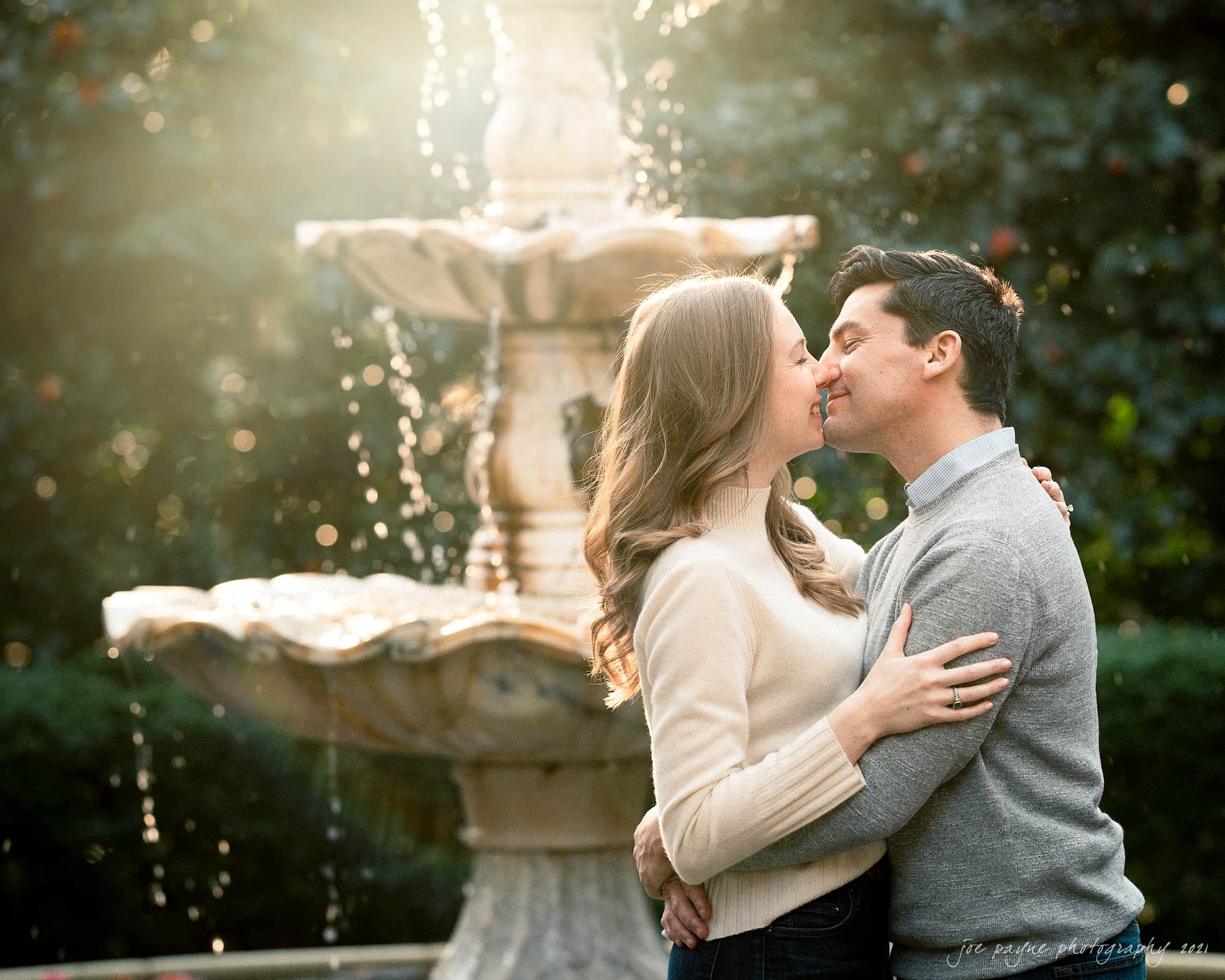 A couple warmly embraces and kisses in front of a flowing fountain. Sunlight filters through, creating a soft glow around them. The background is lush with greenery.