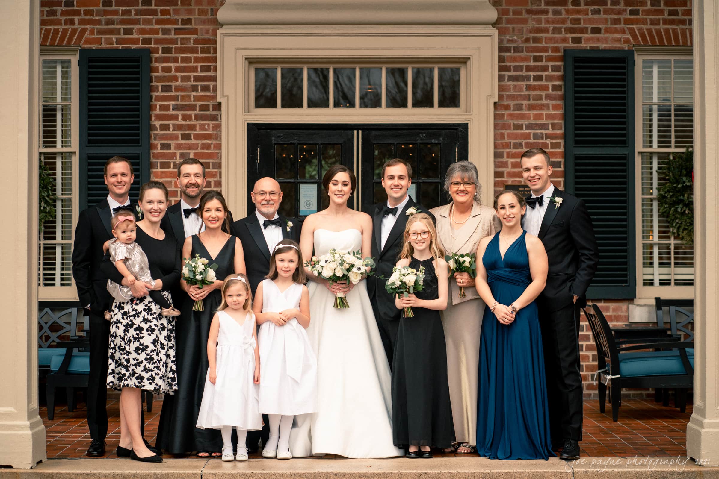 A group wedding photo taken outside the Carolina Inn features the bride and groom at the center, surrounded by family of various ages. The bride wears a white dress, with others in formal attire holding bouquets, highlighting a beautiful Carolina Inn wedding celebration.