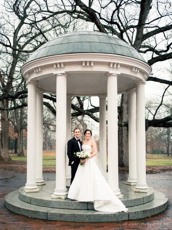 A couple stands under a small, round, white gazebo with columns at their Carolina Inn wedding. The bride is in a strapless white gown, holding a bouquet of white flowers. The groom is in a black suit and tie. The background features bare trees and a brick path.