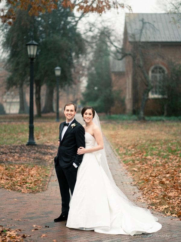 A bride and groom pose happily on a leaf-strewn path at their Carolina Inn wedding. The bride, in a white gown and veil, stands behind the groom in his black tuxedo. They are surrounded by foggy trees and historic buildings on an overcast day.