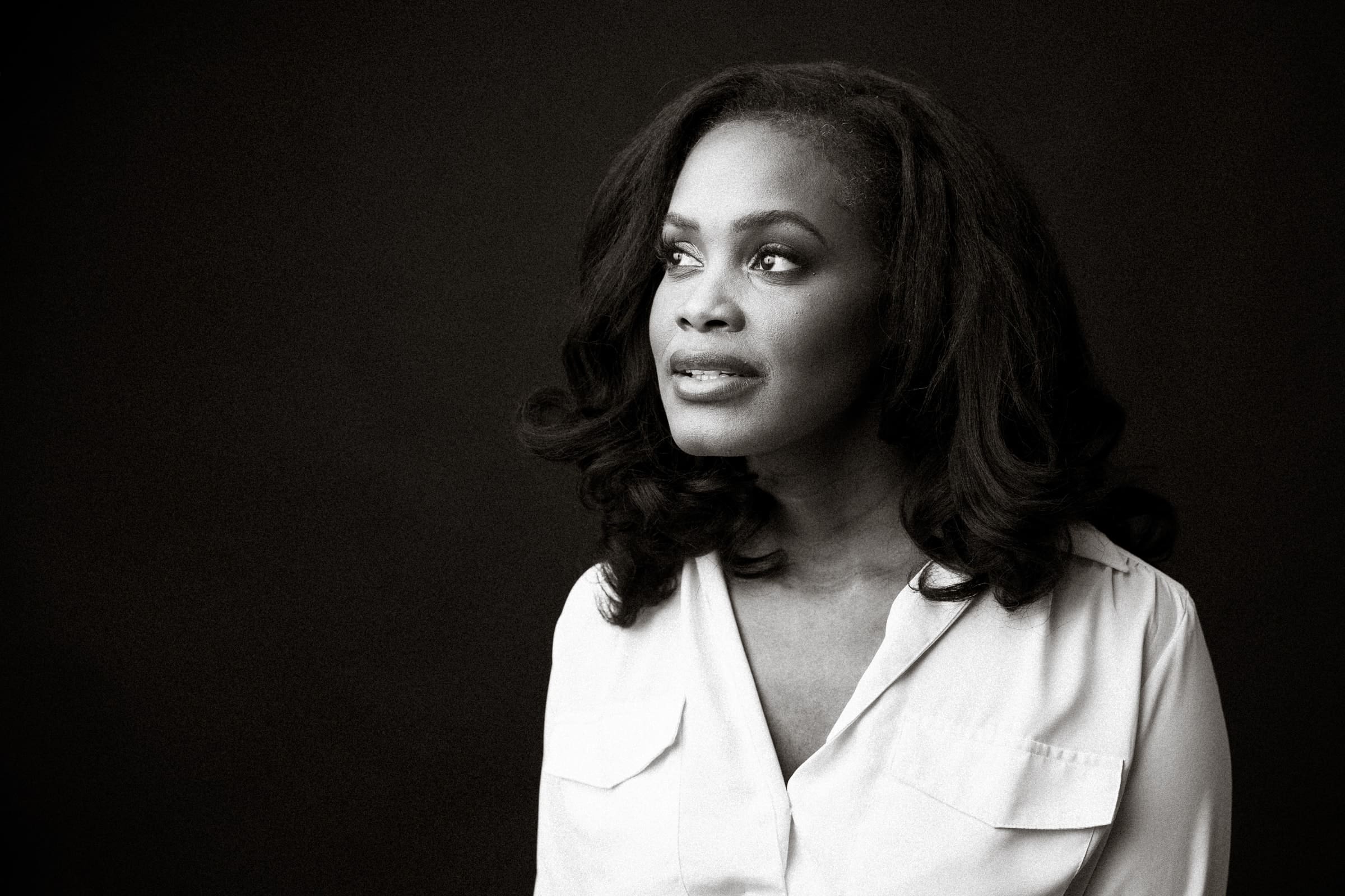 A black and white portrait of a person looking to the side, wearing a button-up shirt, with shoulder-length wavy hair, against a dark background.