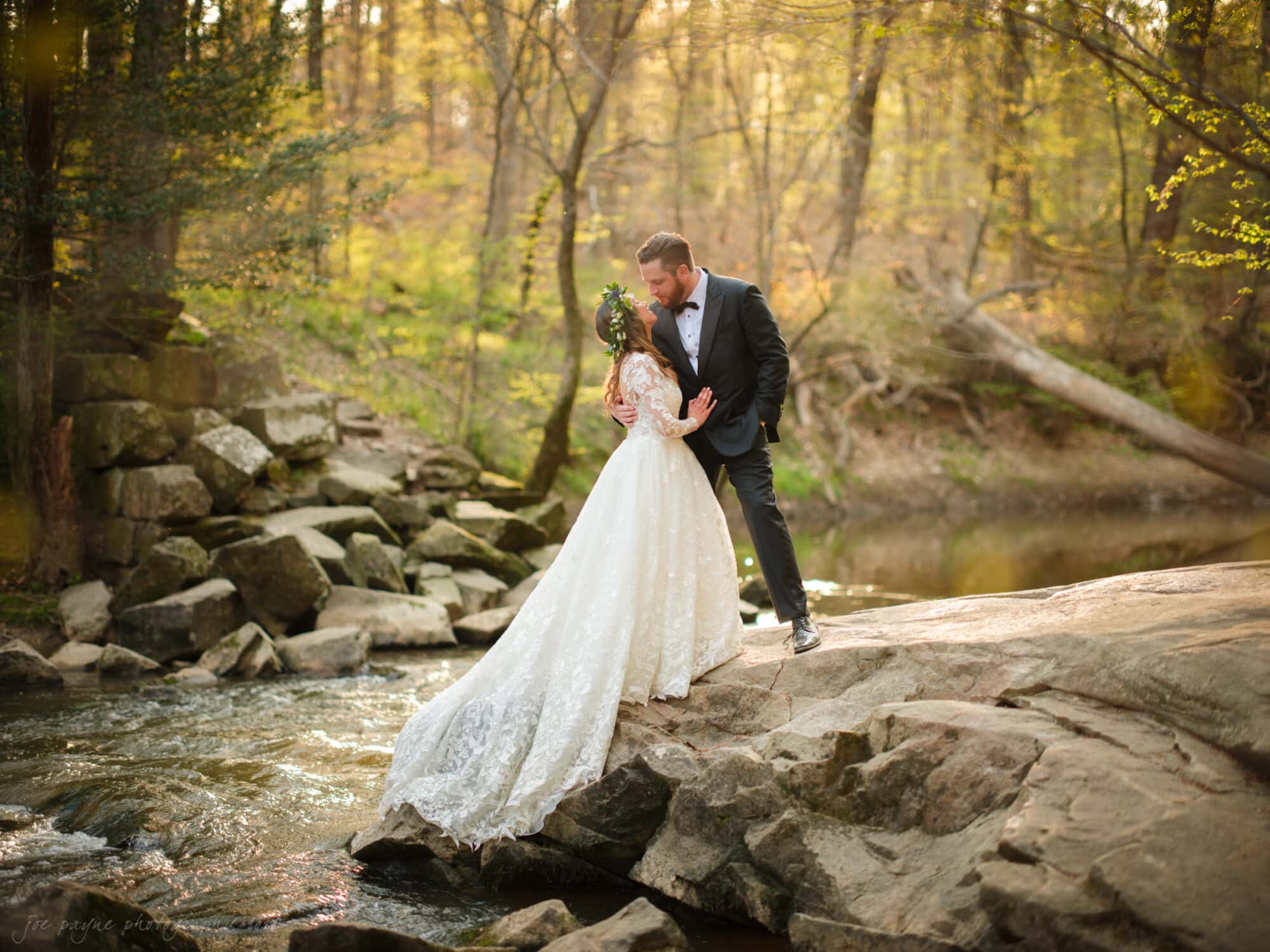 A couple in wedding attire embraces on a rocky edge beside a flowing stream, surrounded by the sunlit Umstead Hotel wedding grounds. The bride wears a flowing white gown, and the groom is in a black suit.