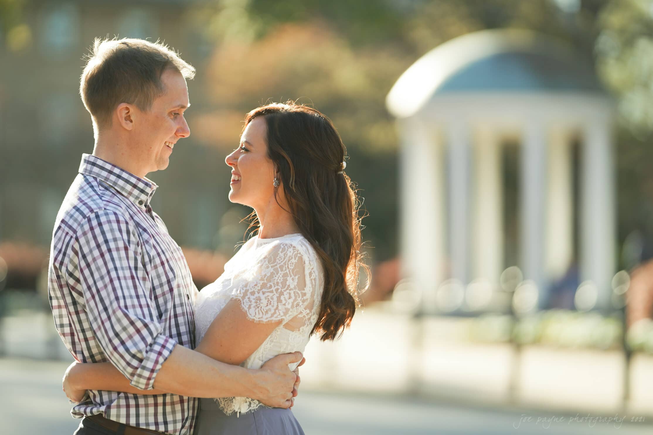 A couple stands close, embracing and smiling at each other in a sunlit outdoor setting, capturing the essence of UNC Chapel Hill engagement photography. The background features a blurred, domed structure, adding to the warm and romantic atmosphere.