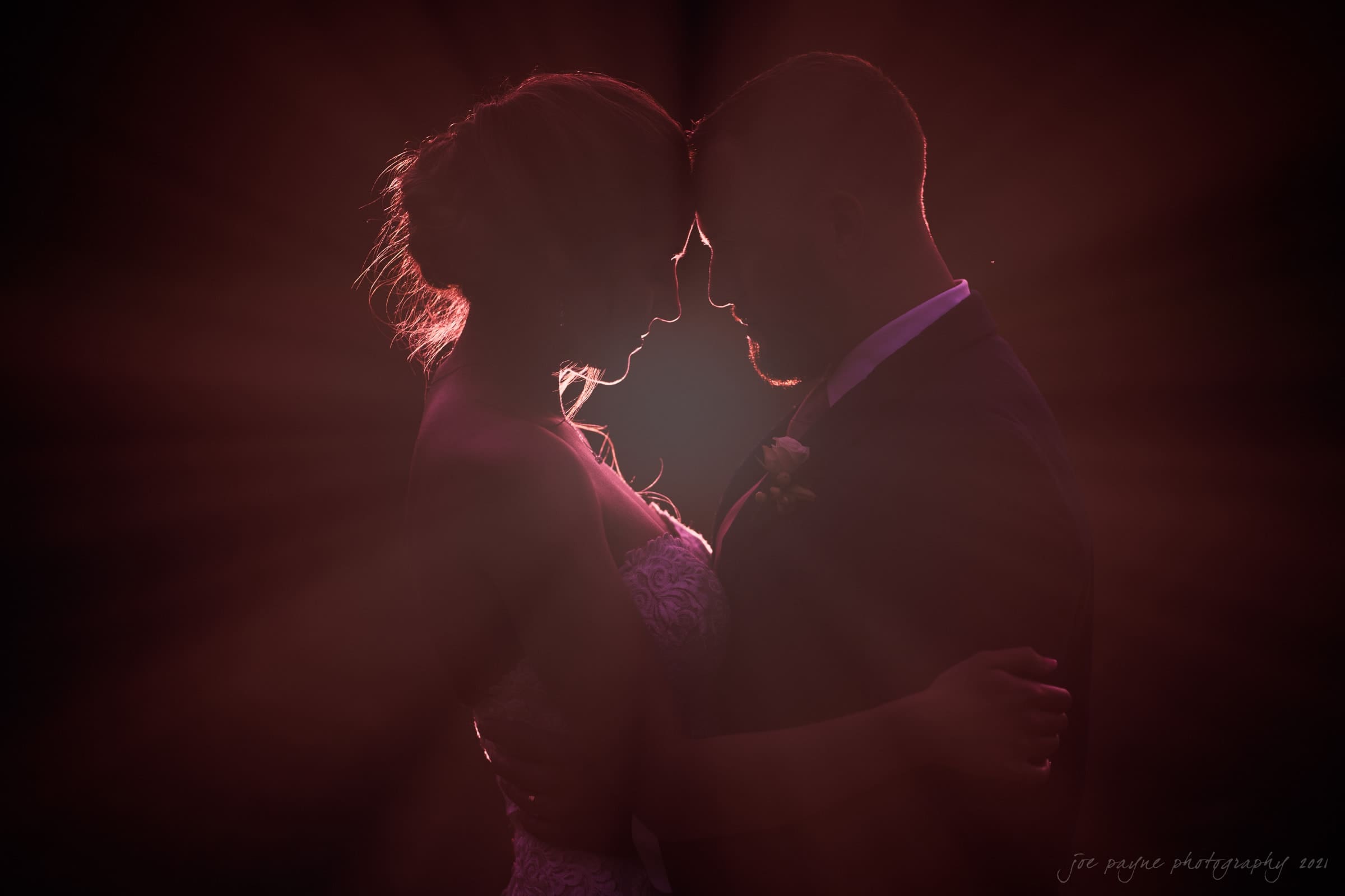 A silhouette of a couple embracing closely, their foreheads touching, exudes warmth and romance. Bathed in ethereal glow, this tender moment feels like a scene from a Sacred Heart Church ceremony or a Pavilion at Carriage Farm wedding, capturing the essence of love.