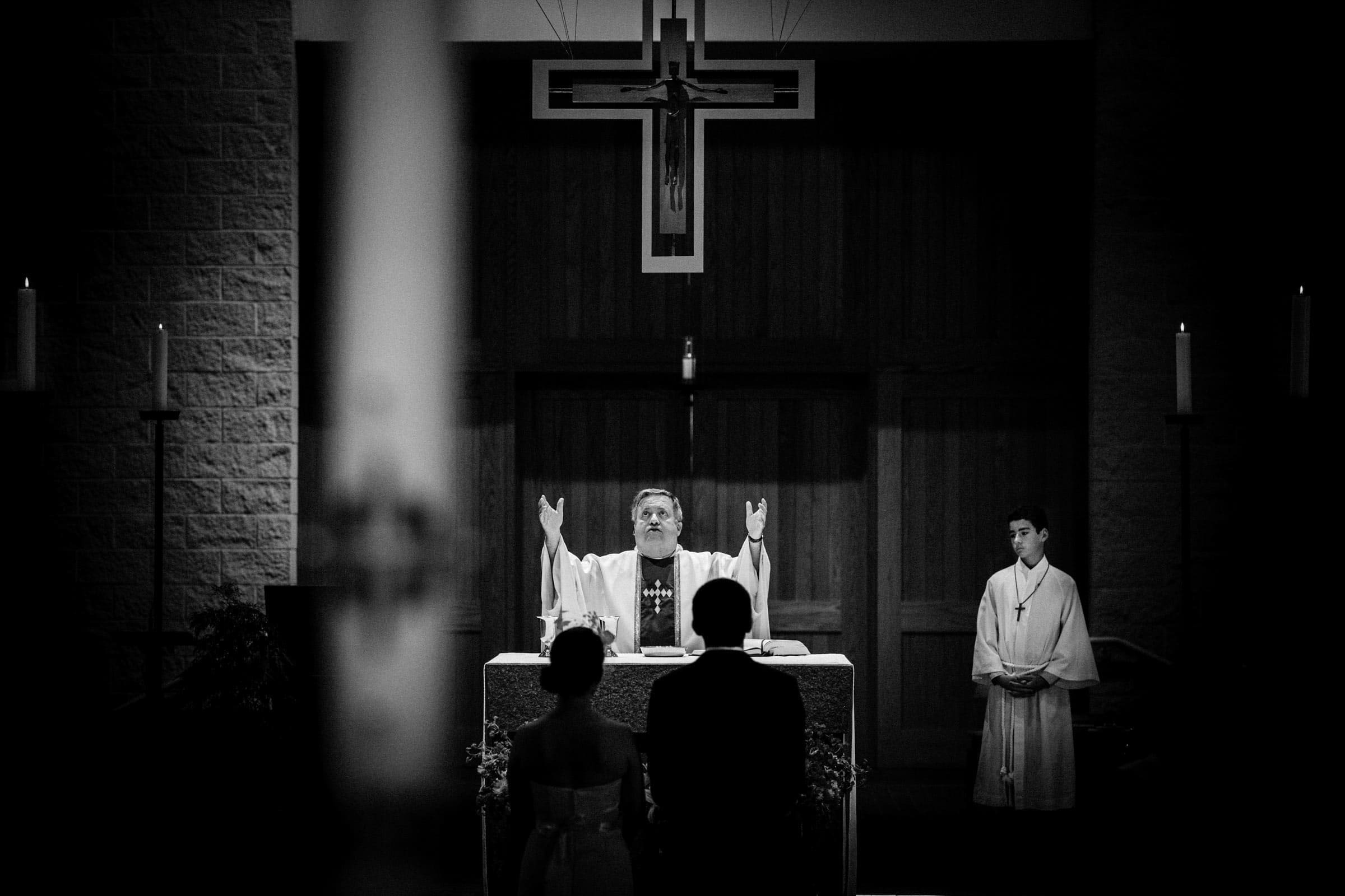 A priest with raised arms stands at an altar beneath a cross, officiating a wedding ceremony for a couple facing him, while an altar server stands to the side. This black and white moment captures the essence of Wedding Photojournalism.