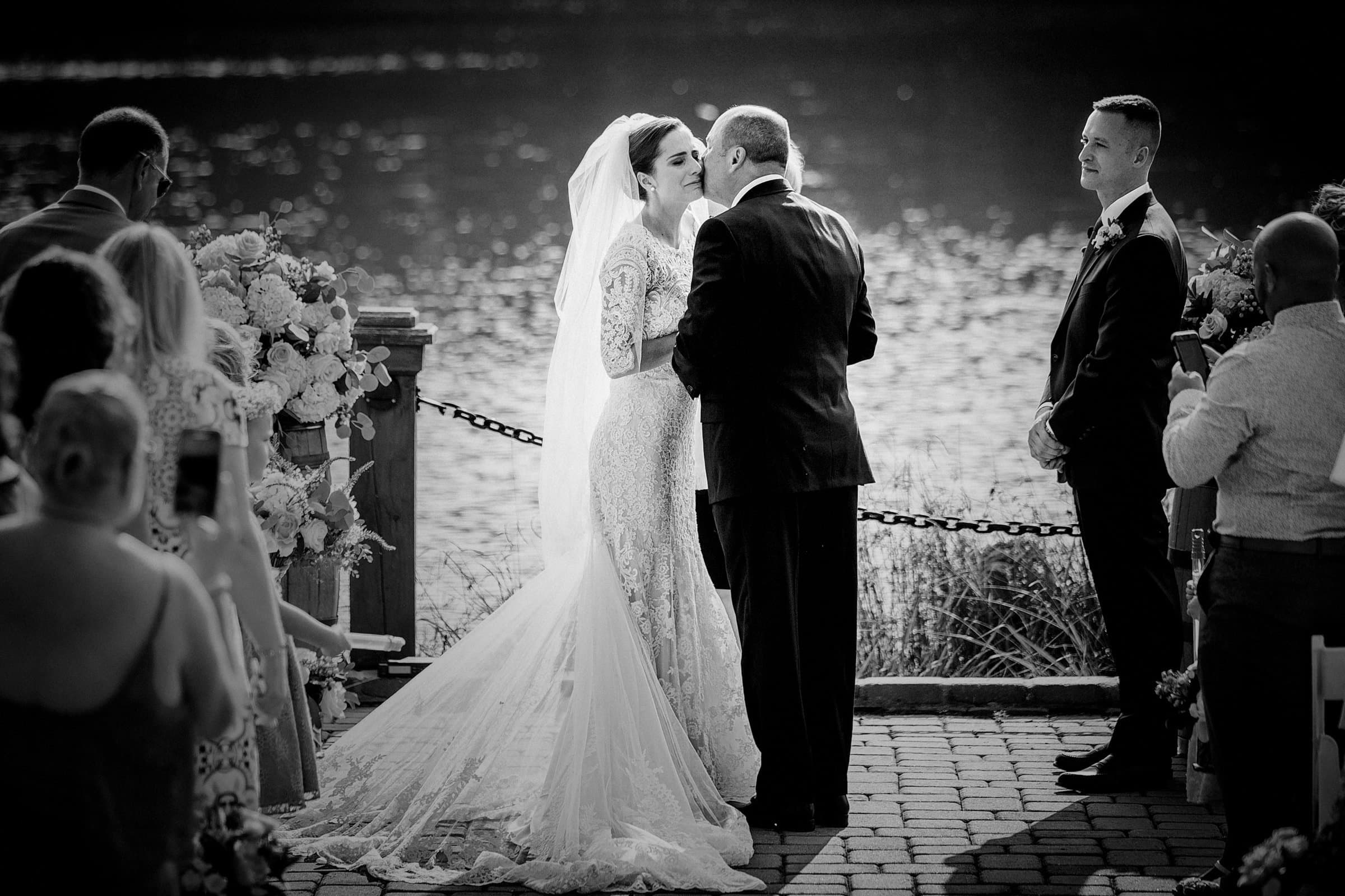 A bride in a lace gown and veil is kissed on the cheek by her father beside a lake during an outdoor Pavilion at Carriage Farm wedding, while the groom and guests look on.