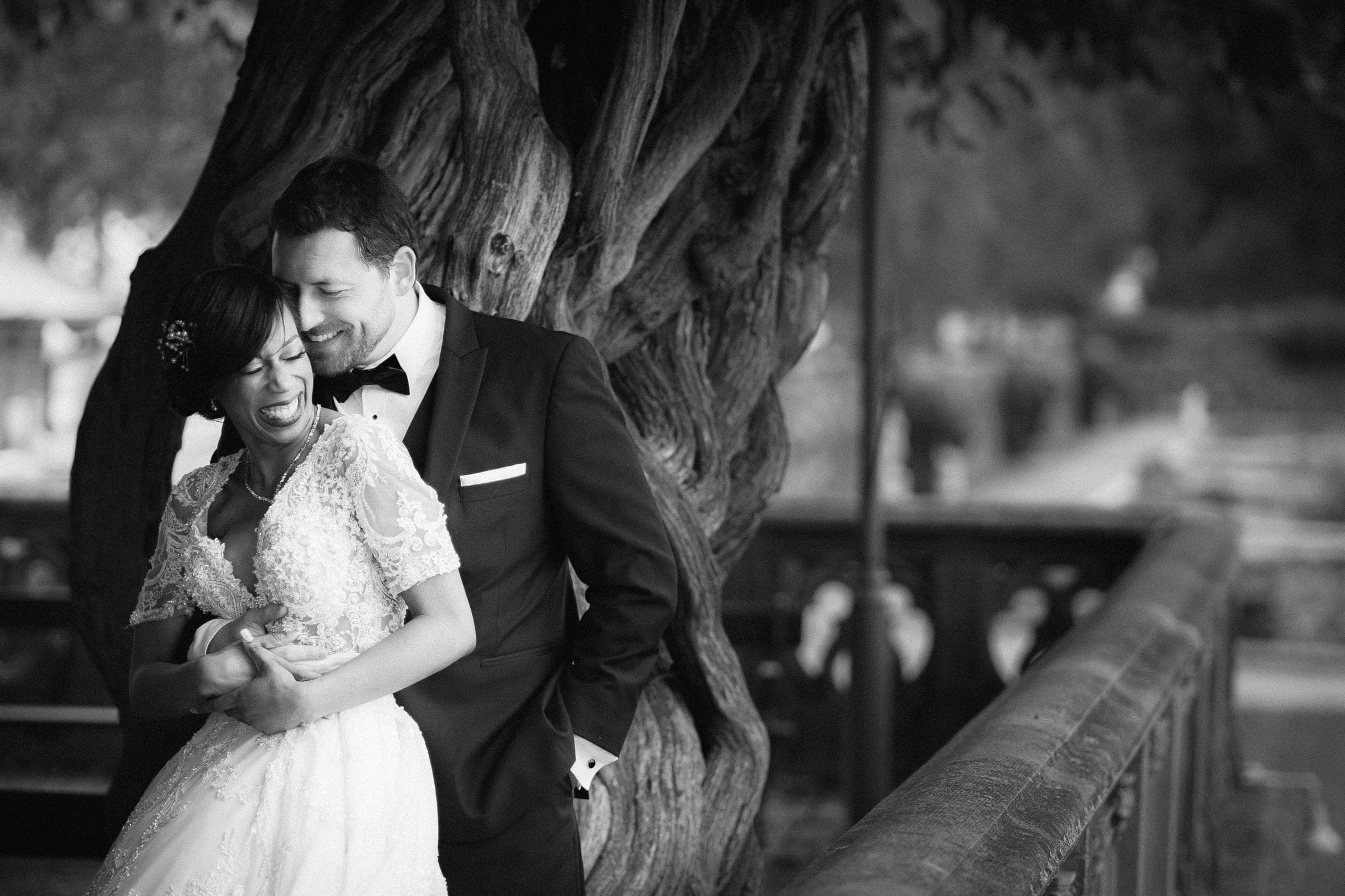 A black-and-white photo of a bride and groom laughing together, captured by a Raleigh photographer. The groom, in a suit, embraces the smiling bride in her lace wedding dress. They stand before a large, twisted tree with a blurred background, epitomizing the charm of Raleigh weddings.