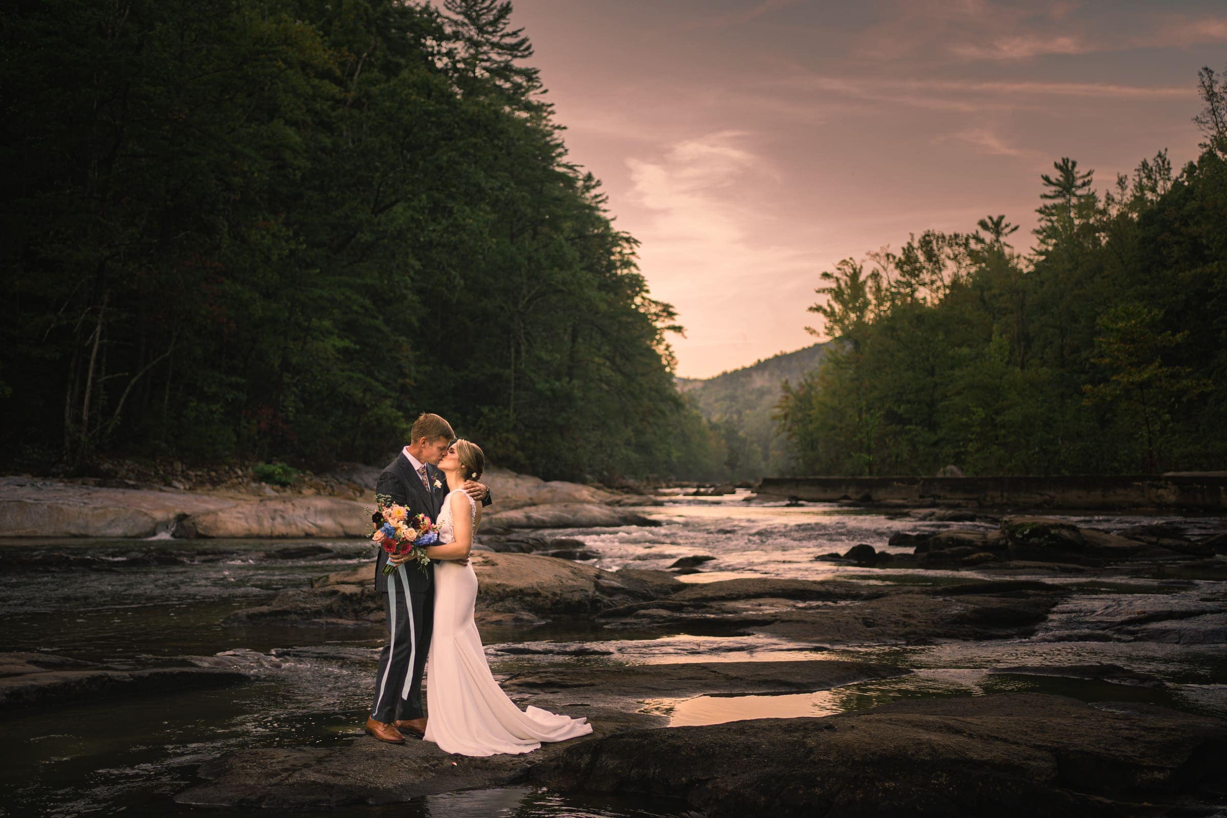 A couple embraces on a rocky riverbank at sunset near Raleigh, surrounded by a forest. The woman holds a colorful bouquet. The sky is a warm pink, adding a romantic atmosphere to the scene captured beautifully by local Durham and Chapel Hill wedding photographers.