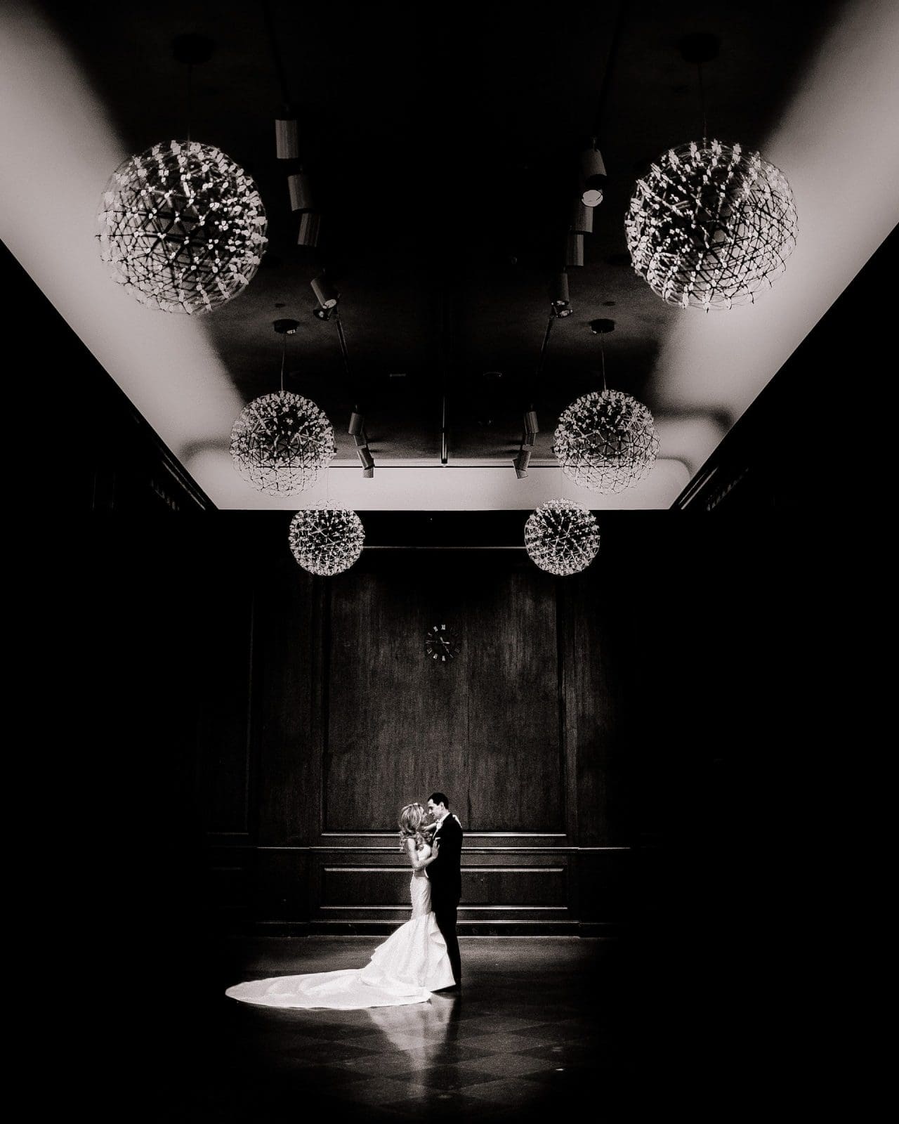 In a dimly lit room with a high ceiling, a couple shares an intimate dance. Spherical chandeliers cast a soft glow over the brides long train trailing elegantly behind her. This enchanting Raleigh setting perfectly captures the essence cherished by wedding and portrait photographers.
