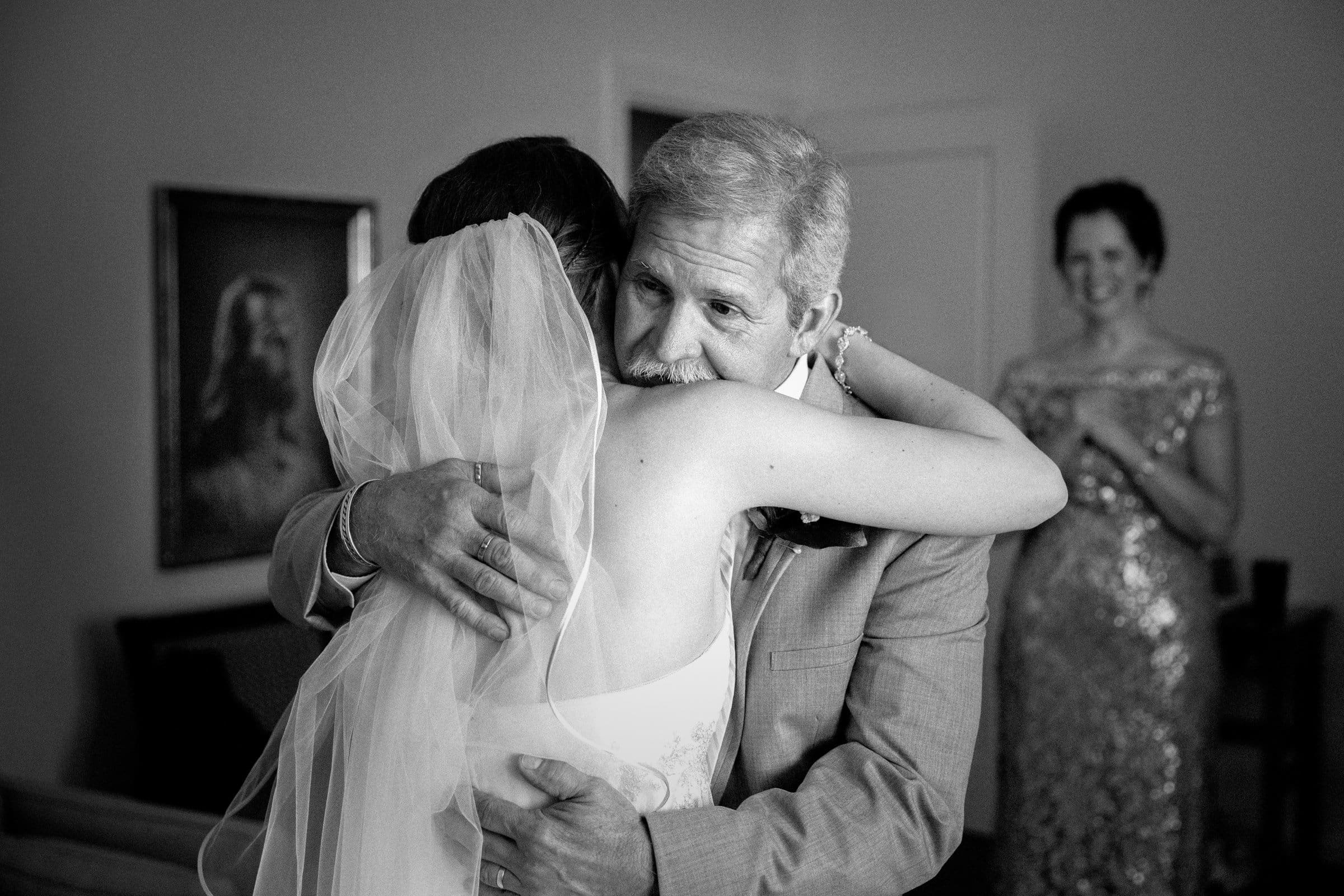 A bride in a veil hugs an older man, likely her father, while a woman in a sparkling dress smiles warmly in the background. The scene appears emotional and joyful, likely moments before a wedding ceremony.