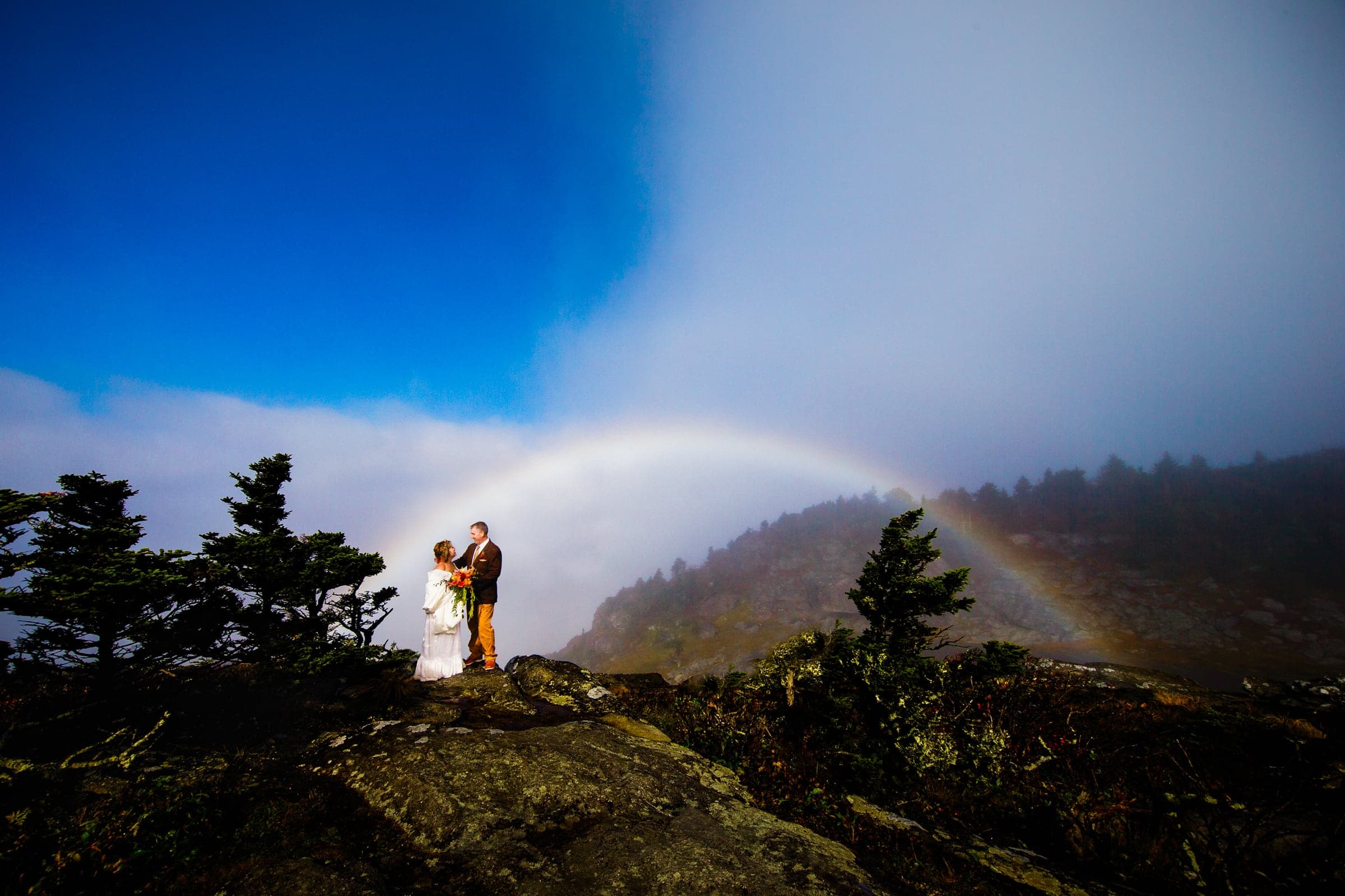 A couple stands on a rocky mountain peak, embracing under a colorful rainbow. The sky is partially blue with some mist and clouds. Evergreen trees and the mountain landscape set the scene, reminiscent of locations favored by top Raleigh photographers for their stunning wedding portraits.