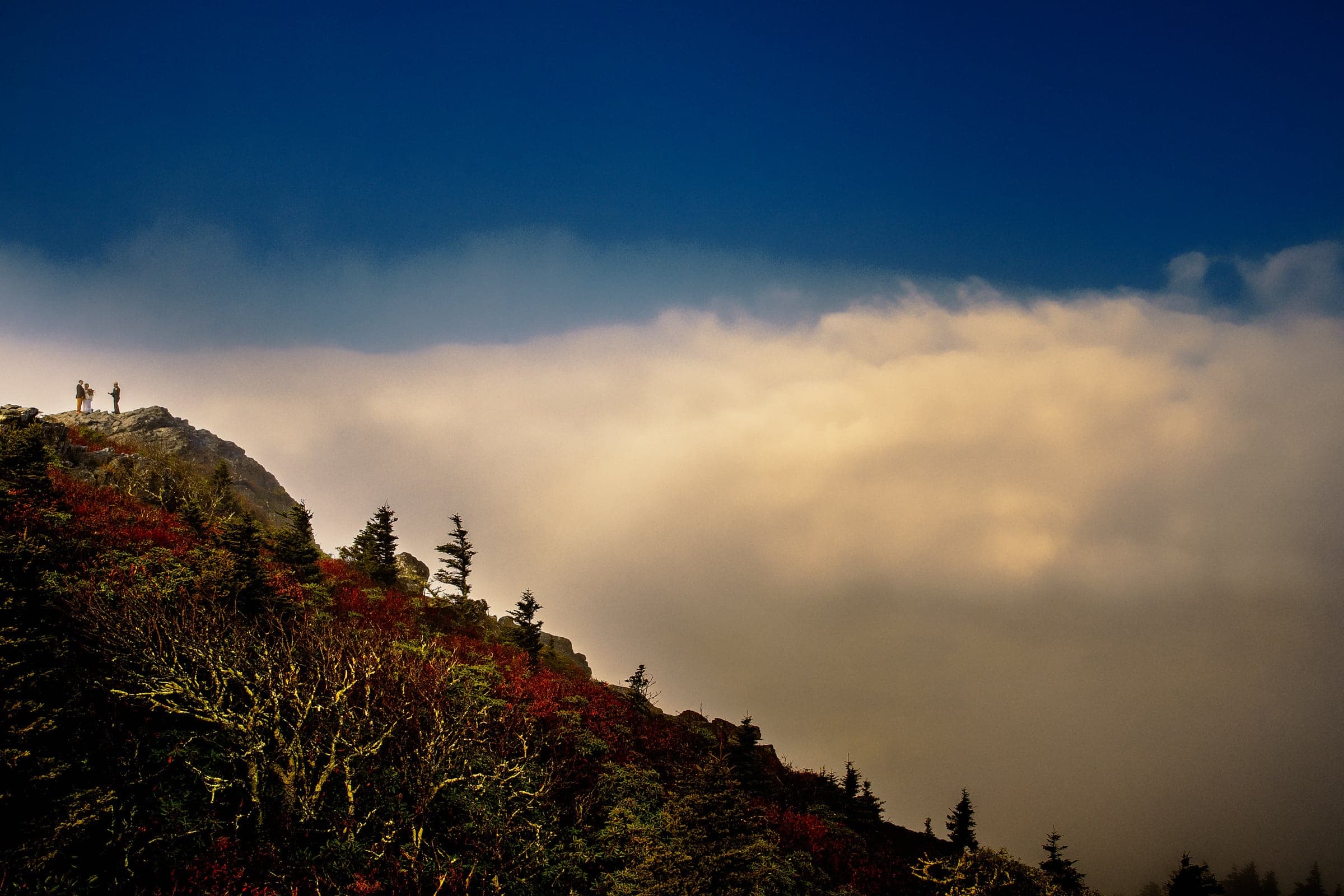 Two people stand on a rocky peak overlooking a landscape with dense clouds and a clear blue sky, reminiscent of the stunning vistas Raleigh offers. Trees in shades of red and orange autumn foliage frame this perfect moment, ideal for any wedding or portrait by Durham & Chapel Hill's top photographers.