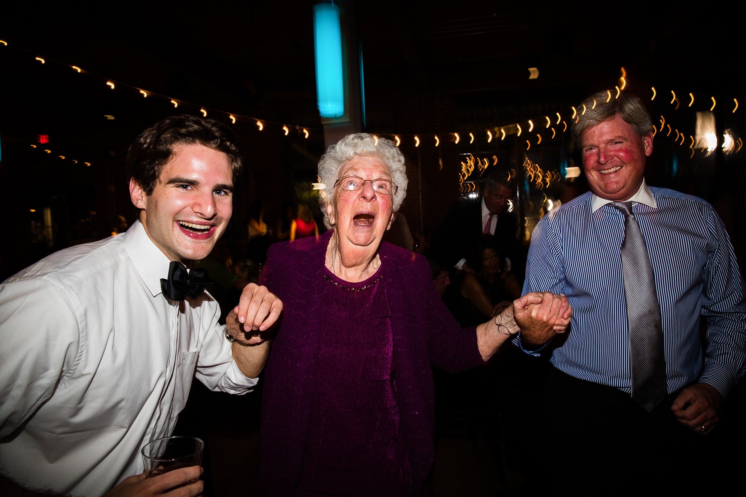 An elderly woman in a purple outfit laughs joyfully while holding hands with two men in dress shirts and ties, perfectly capturing the candid spirit of wedding photojournalism at a lively indoor celebration with glowing string lights.