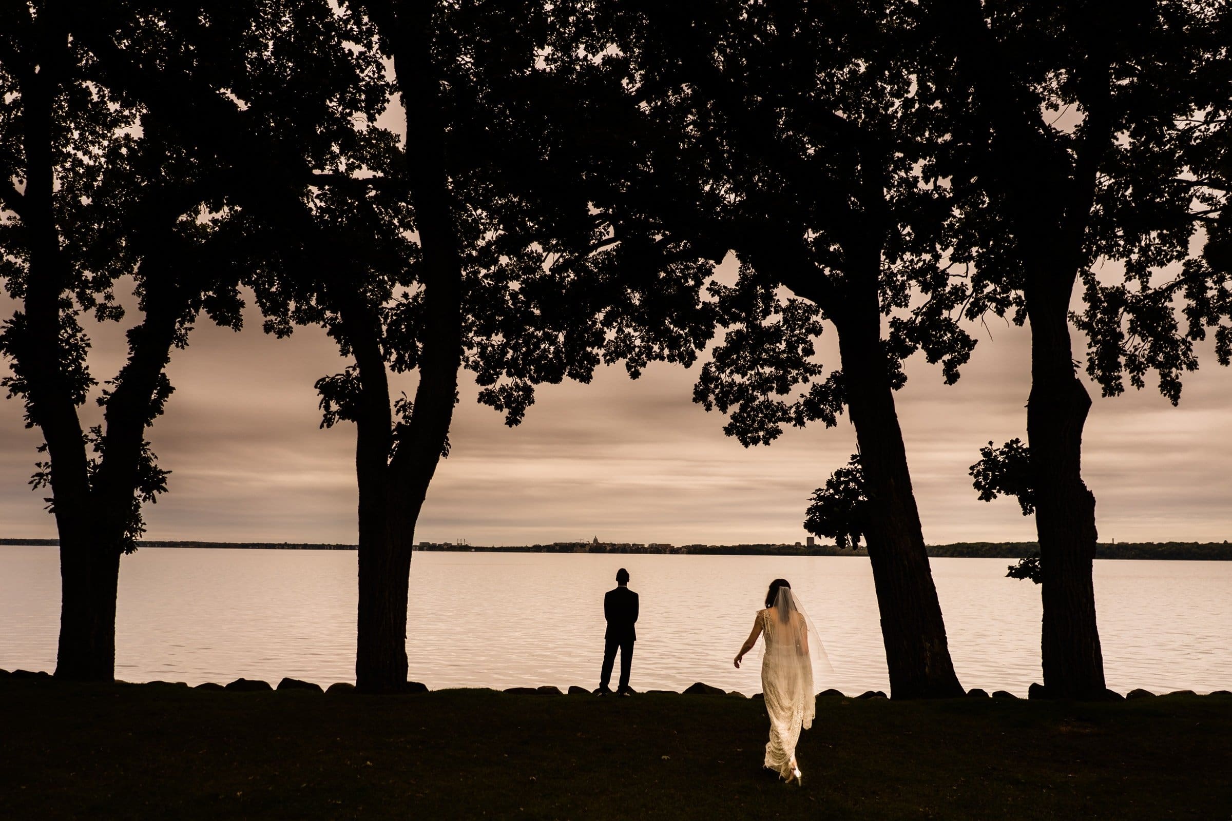 A bride in a flowing dress walks toward a groom standing by a tranquil Raleigh lake, framed by silhouetted trees against a moody, overcast sky—a perfect scene captured by Durham & Chapel Hill wedding photographers.