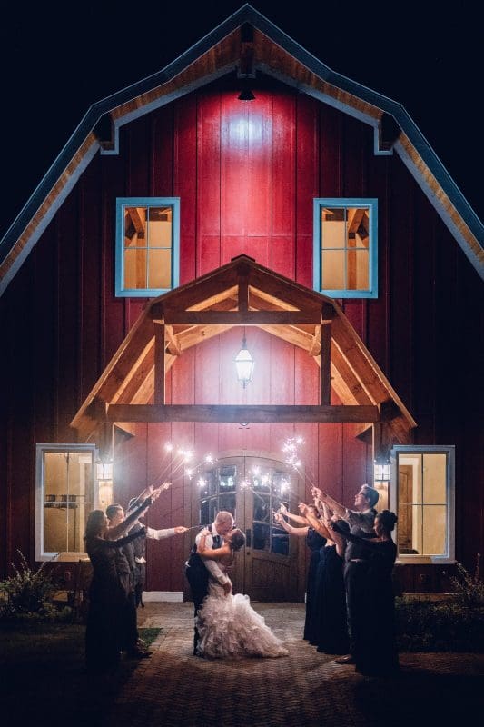 A bride and groom share a tender kiss under a canopy of sparklers held by guests at this stunning Raleigh celebration. They stand in front of a large red barn, warmly lit from inside, creating a romantic evening scene perfect for any Durham & Chapel Hill wedding photographer’s portfolio.