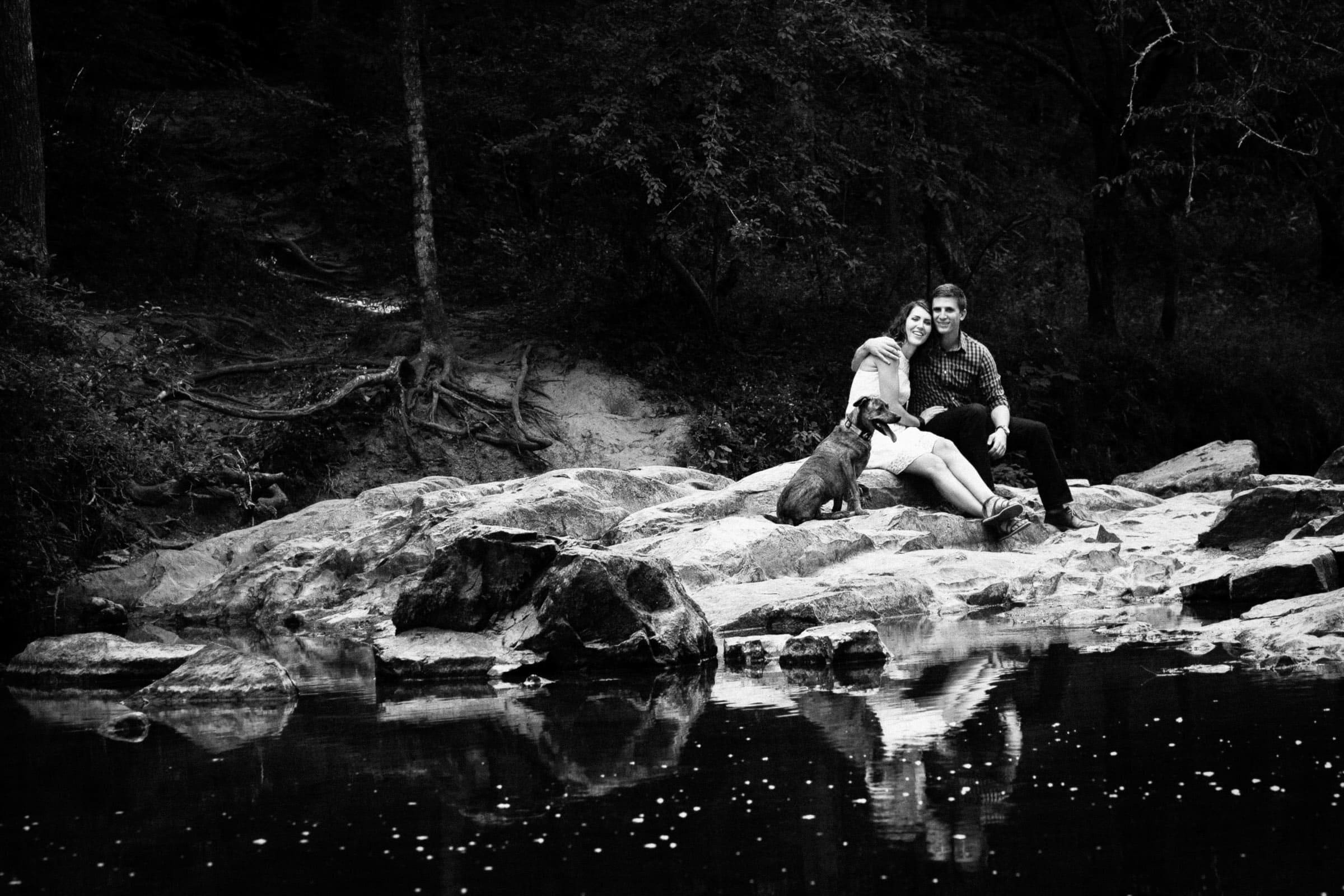 A black and white photo captures a couple during their engagement photo session, sitting on rocks by a calm river, with trees in the background. They are smiling with their dog by their side, who gazes at the water. The serene scene is beautifully reflected in the gentle ripples.