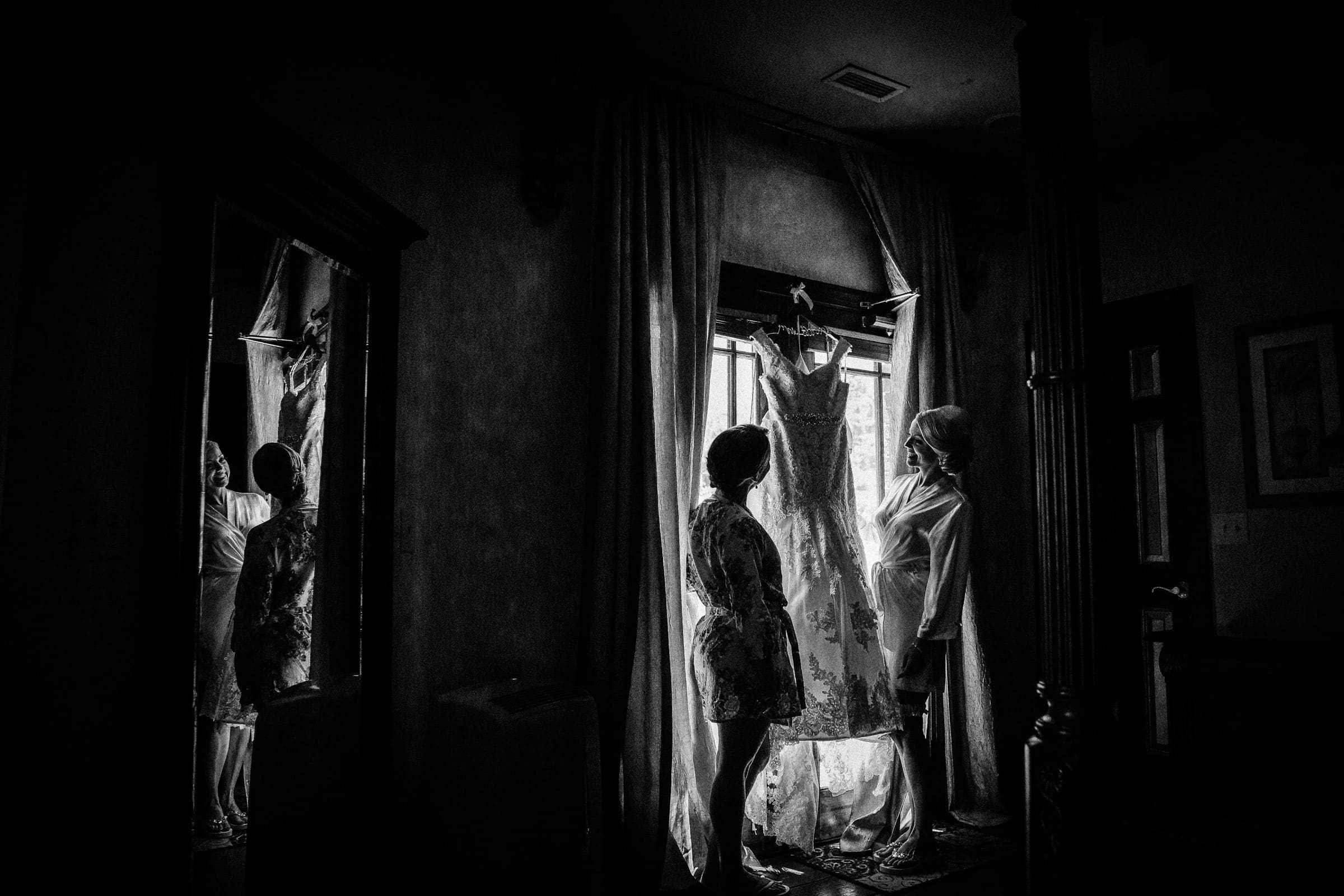 A black and white photo captures two women in robes, one hanging a wedding dress by the window. The serene and intimate scene is beautifully reflected in a nearby mirror, perfectly embodying the elegance often sought after by Raleigh wedding photographers.