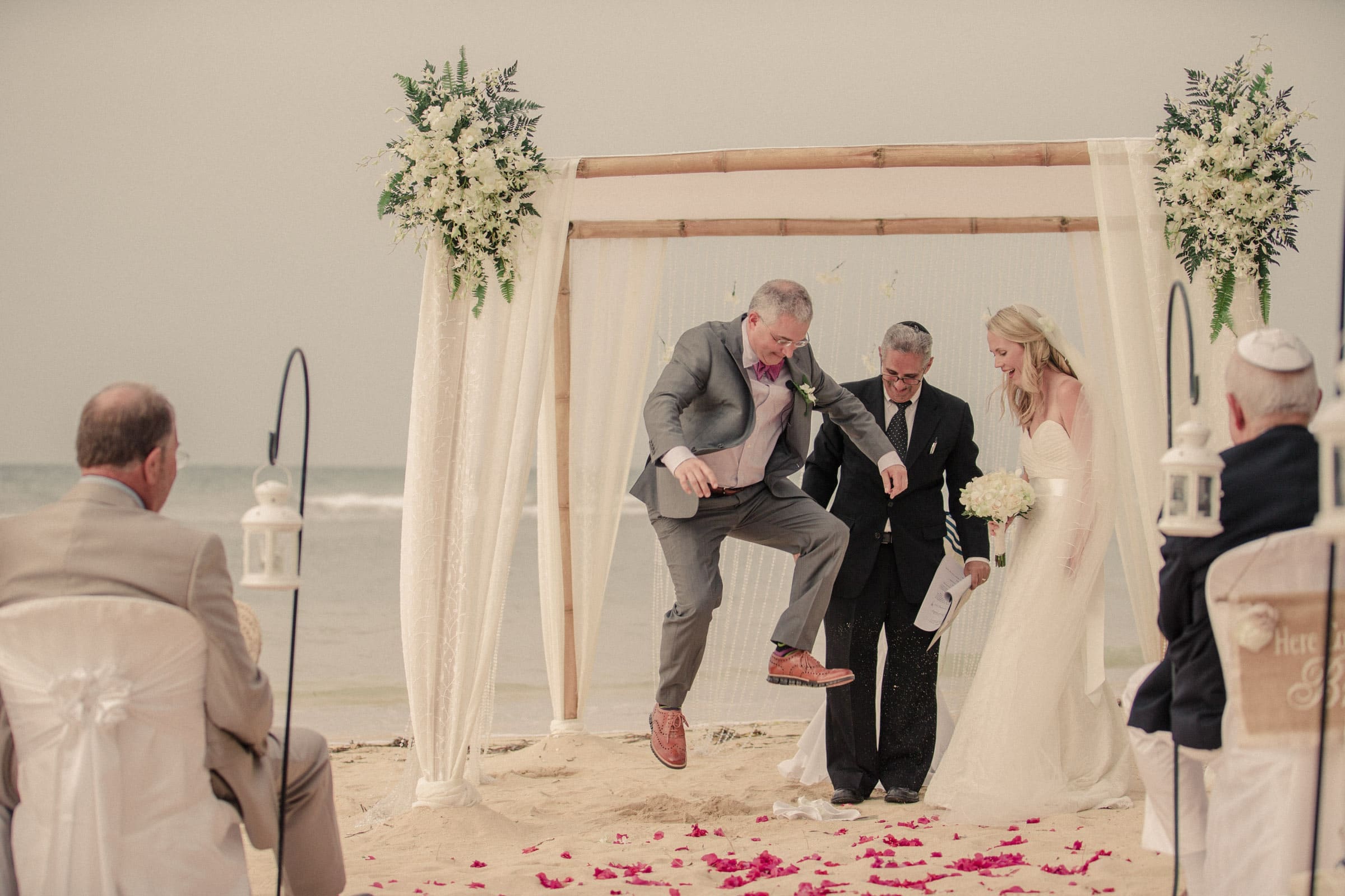 A bride and groom stand under a floral arch on a Raleigh beach. The groom jumps enthusiastically, surrounded by guests on chairs adorned with white covers. A layer of red flower petals covers the sand, perfectly capturing the love and joy that Durham & Chapel Hill wedding photographers seek to frame.