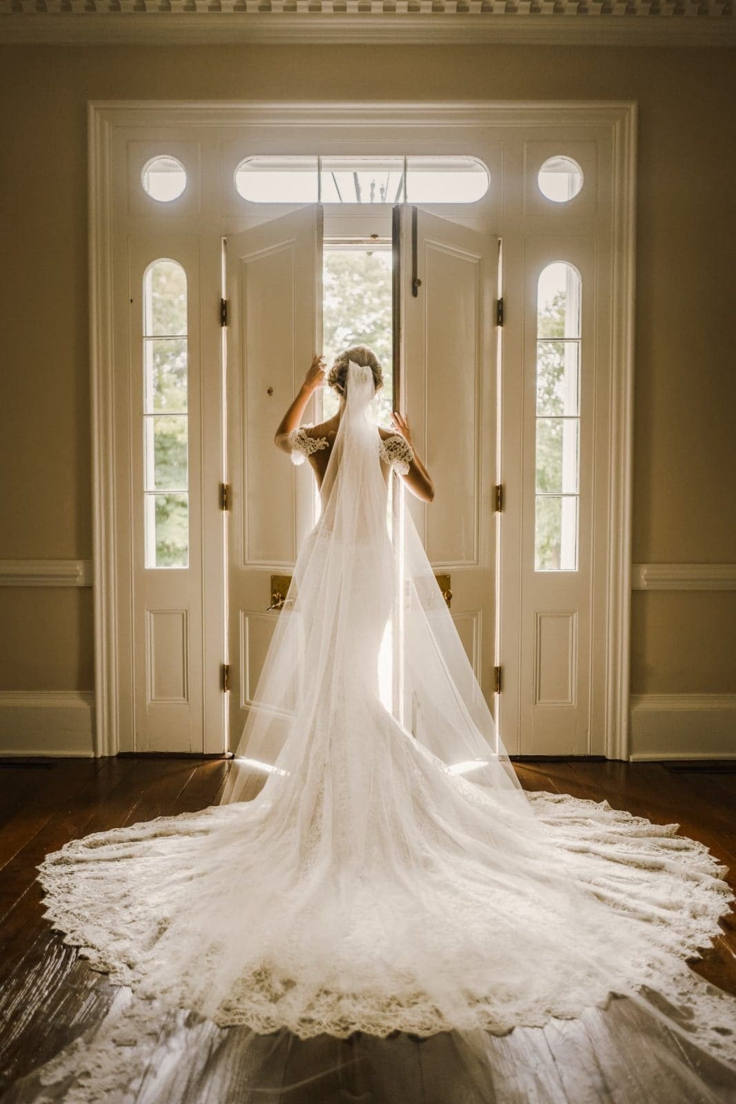 A bride in a white lace wedding dress stands facing an open door. Sunlight streams in, illuminating her veil and the long train of her gown as she preps for her bridal session. The room features elegant wood flooring and decorative molding around the door, offering a perfect setting.