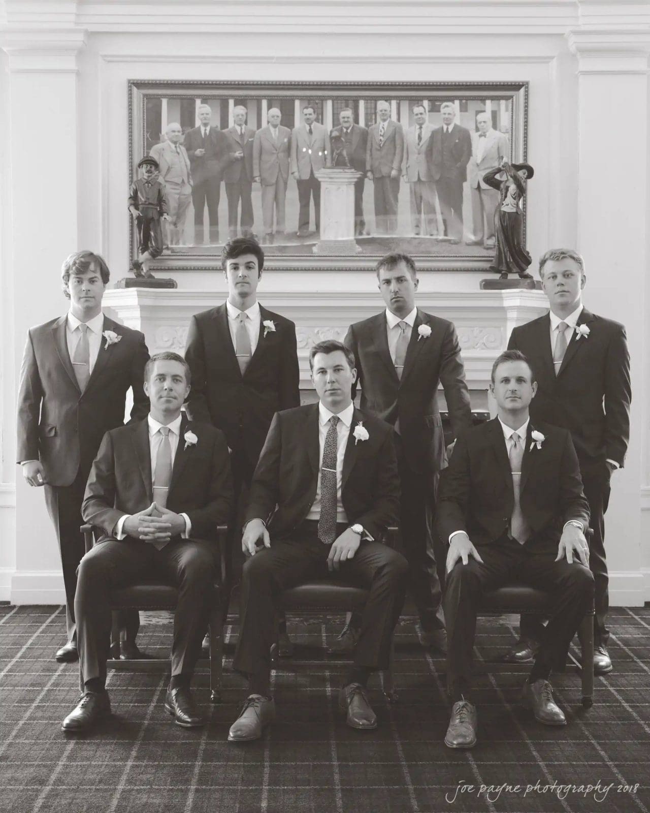 A group of seven men in suits poses for a formal photograph in a room with a large painting and statues. Four men are standing, and three are sitting. They all wear boutonnieres and look at the camera. The photo is in black and white.