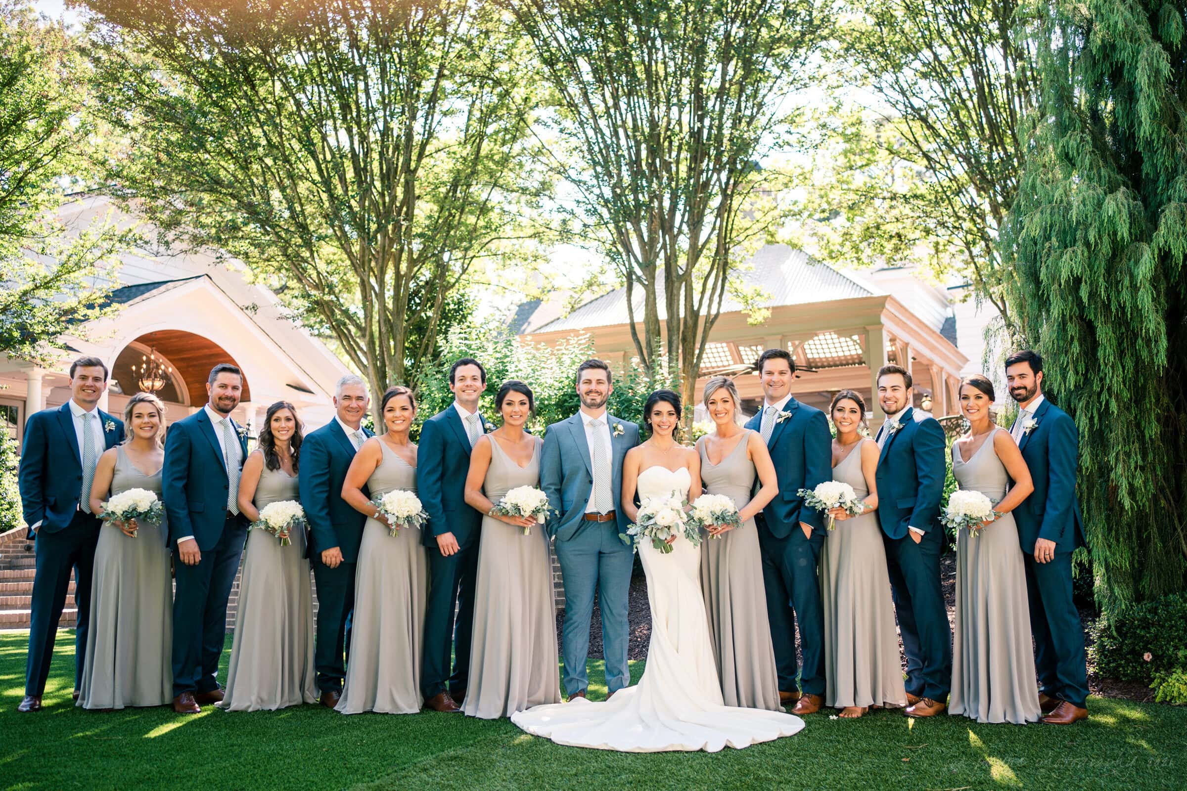 A wedding party poses outdoors at the enchanting Highrove Estate. The bride dazzles in her white gown beside the groom in a light gray suit. Bridesmaids in beige dresses and groomsmen in dark suits smile brightly, holding bouquets, captured beautifully by the talented estate wedding photographer.