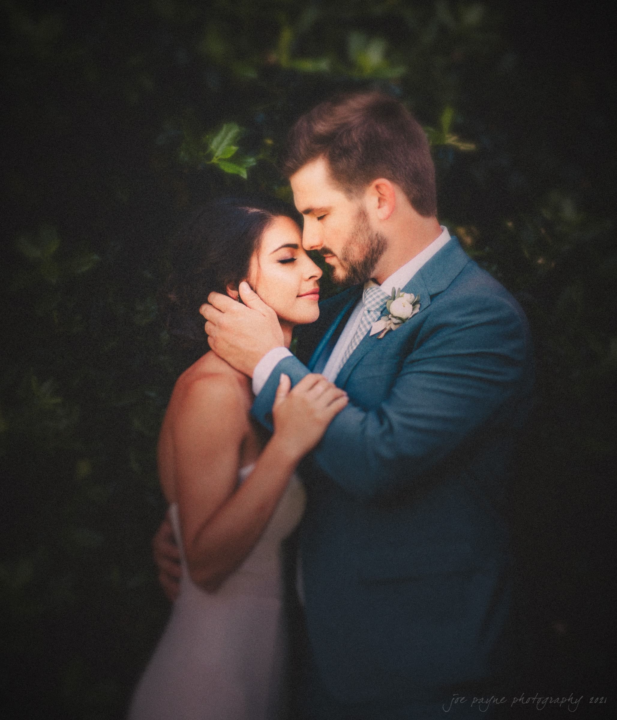 A bride and groom embrace tenderly in front of a lush, green background. The bride wears a strapless white dress, while the groom is in a blue suit with a boutonniere. They appear happy and serene.