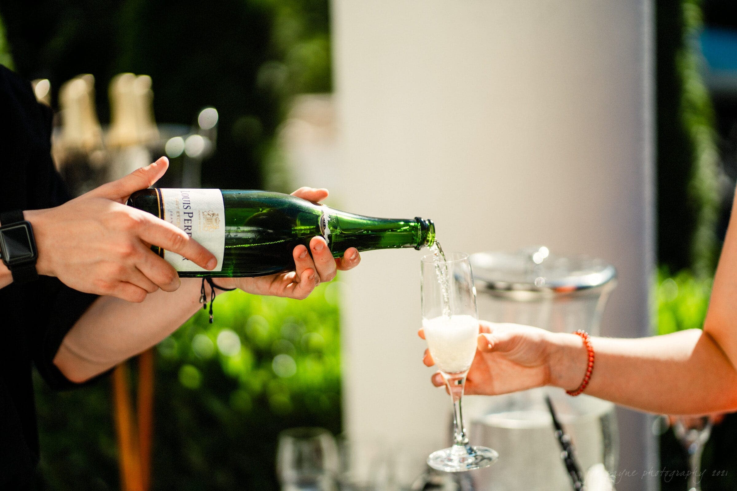 A person pours sparkling wine from a green bottle into a flute held by another person. The scene is outdoors with sunlight, a blurred green background, and a wine bucket nearby.