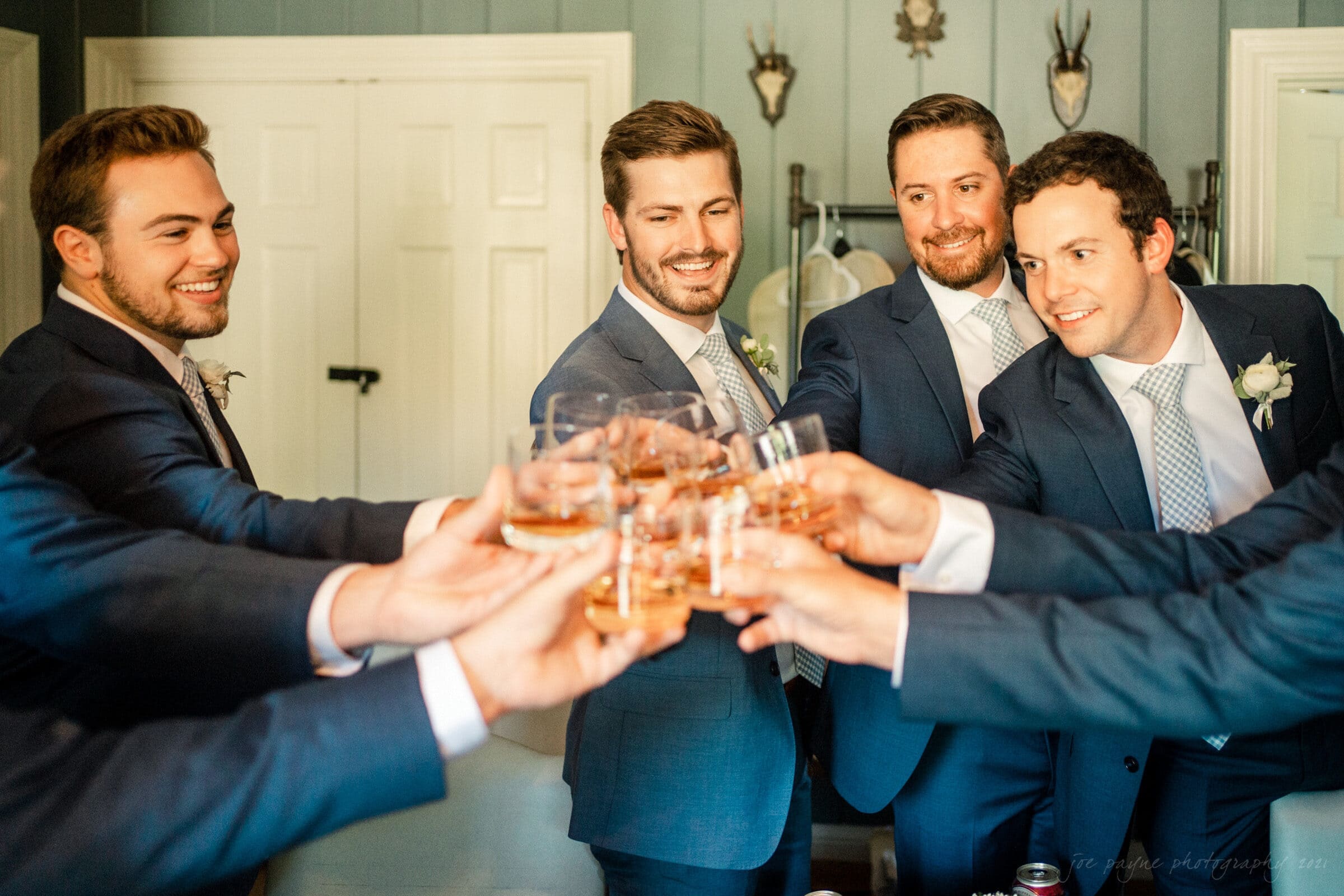 A group of five men in suits clinking glasses in a toast, smiling and standing in a room with closed doors and decorations. They appear to be celebrating a special occasion, like a wedding.