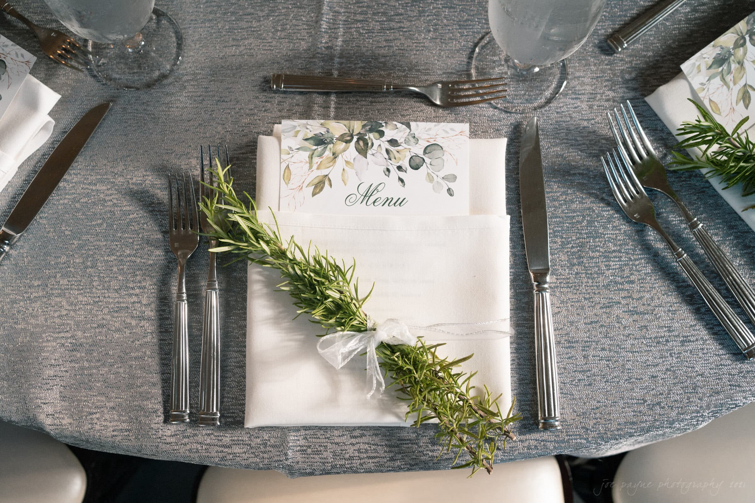 A formal place setting on a gray tablecloth, featuring a white napkin with a sprig of rosemary tied with a ribbon. A menu with a floral design is partially visible. Silverware and clear water glasses complete the elegant arrangement.