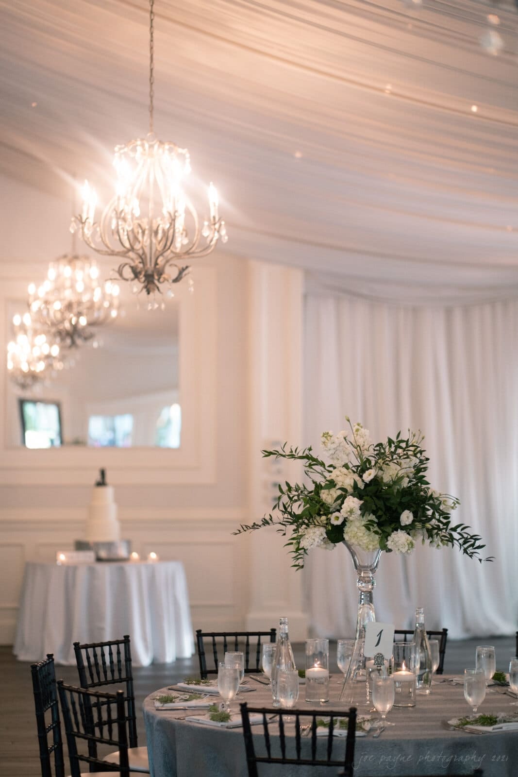 A beautifully decorated event space with a round table set with glassware, silver bottles, and a tall floral centerpiece. Elegant chandeliers hang from a draped ceiling. A white tiered cake is visible in the background.