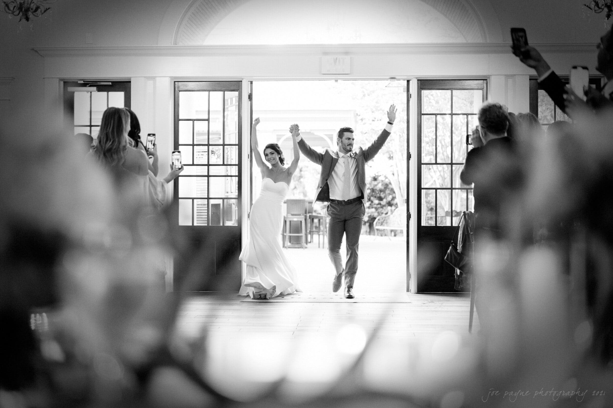A bride and groom joyfully enter a reception hall, hands raised in celebration. Guests stand and take photos with their phones as the couple walks in, with sunlight streaming through the open doors behind them. Black and white image captures the festive moment.