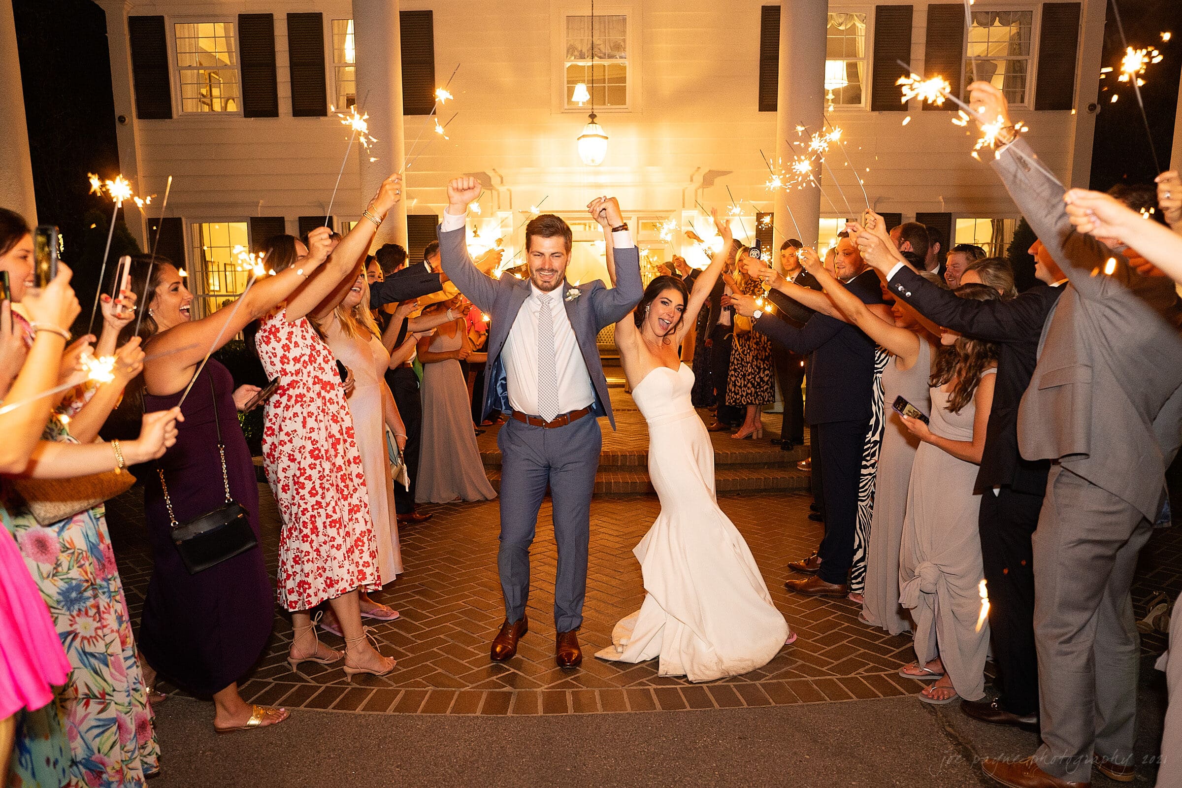A bride and groom joyfully walk under a tunnel of sparklers held by wedding guests outside a large house at night. The bride is in a white gown, and the groom is in a suit, both celebrating with raised arms.