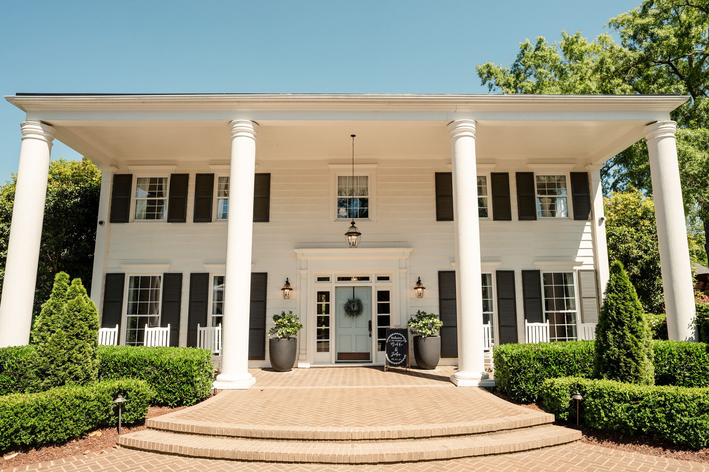 A two-story, white colonial-style house with tall columns and black shutters, perfect for capturing timeless images by a Highrove Estate wedding photographer. Its central entrance is adorned with a wreath on the door, flanked by rocking chairs and potted plants amidst neatly trimmed bushes and a brick pathway.
