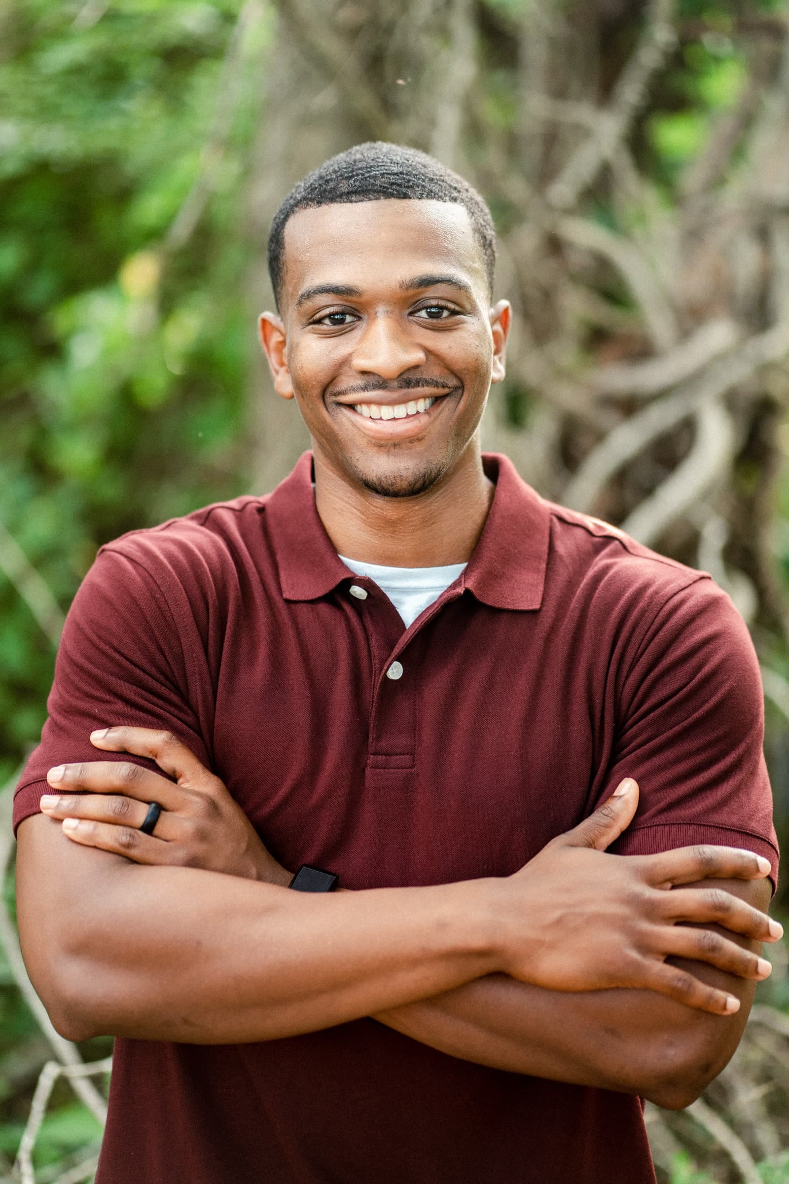 raleigh engagement photographer - kayla & deandre A person wearing a maroon polo shirt smiles with arms crossed. They are standing outdoors with green foliage in the background.