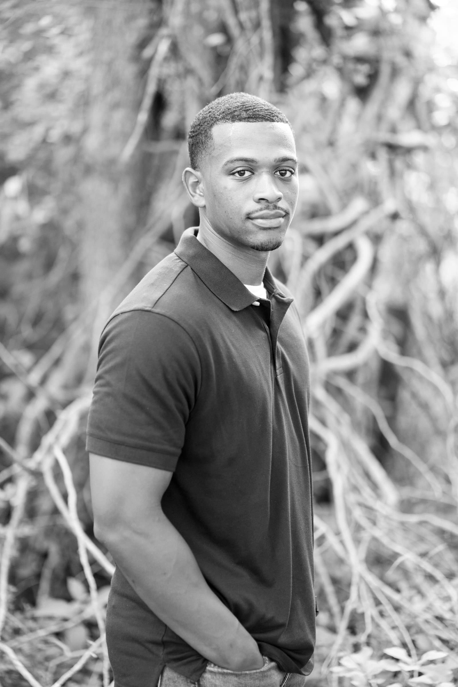 raleigh engagement photographer - kayla & deandre A man stands outdoors in front of a backdrop of intertwined branches and foliage. He is wearing a dark polo shirt and has his hands in his pockets, looking toward the camera with a neutral expression. The image is in black and white.