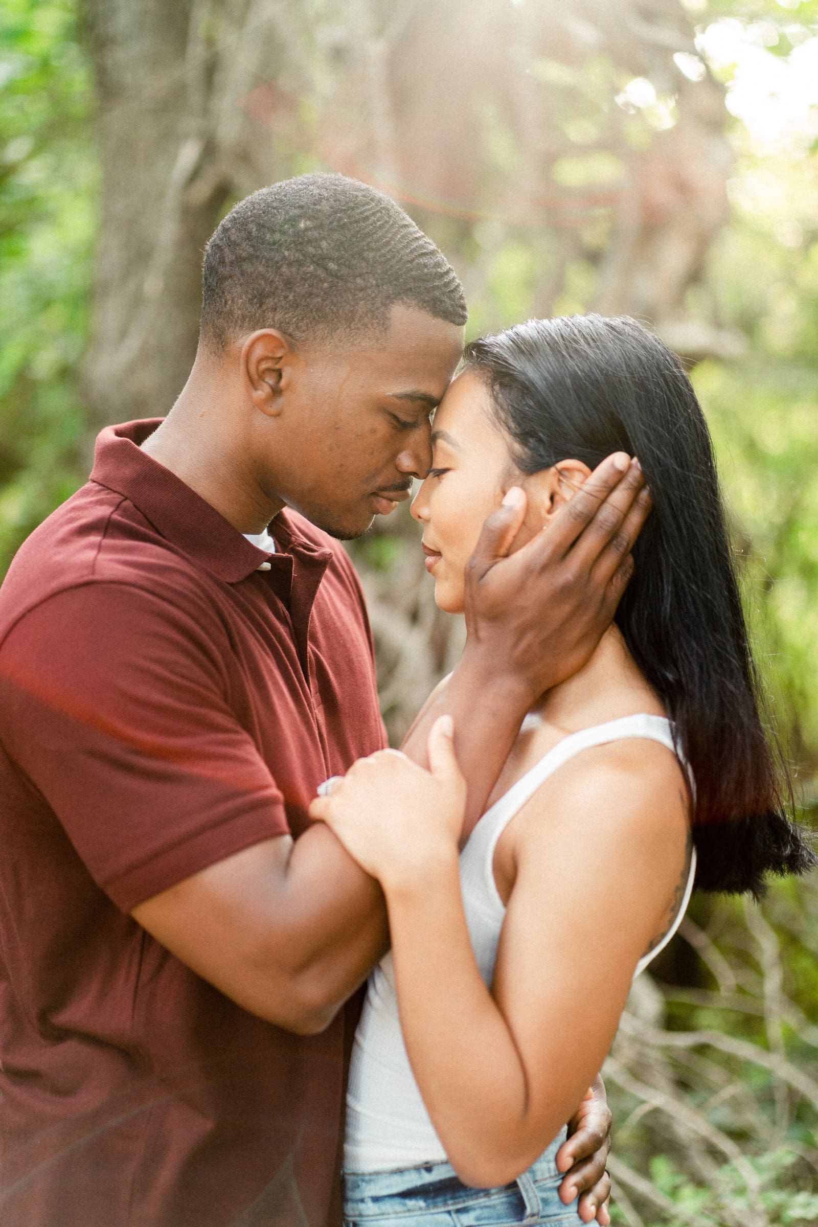 raleigh engagement photographer - kayla & deandre A couple embraces tenderly in a sunlit forest. The man, in a maroon shirt, gently holds the womans face as they lean in close, eyes closed. The woman wears a white tank top, and the scene is peaceful and intimate, surrounded by greenery.