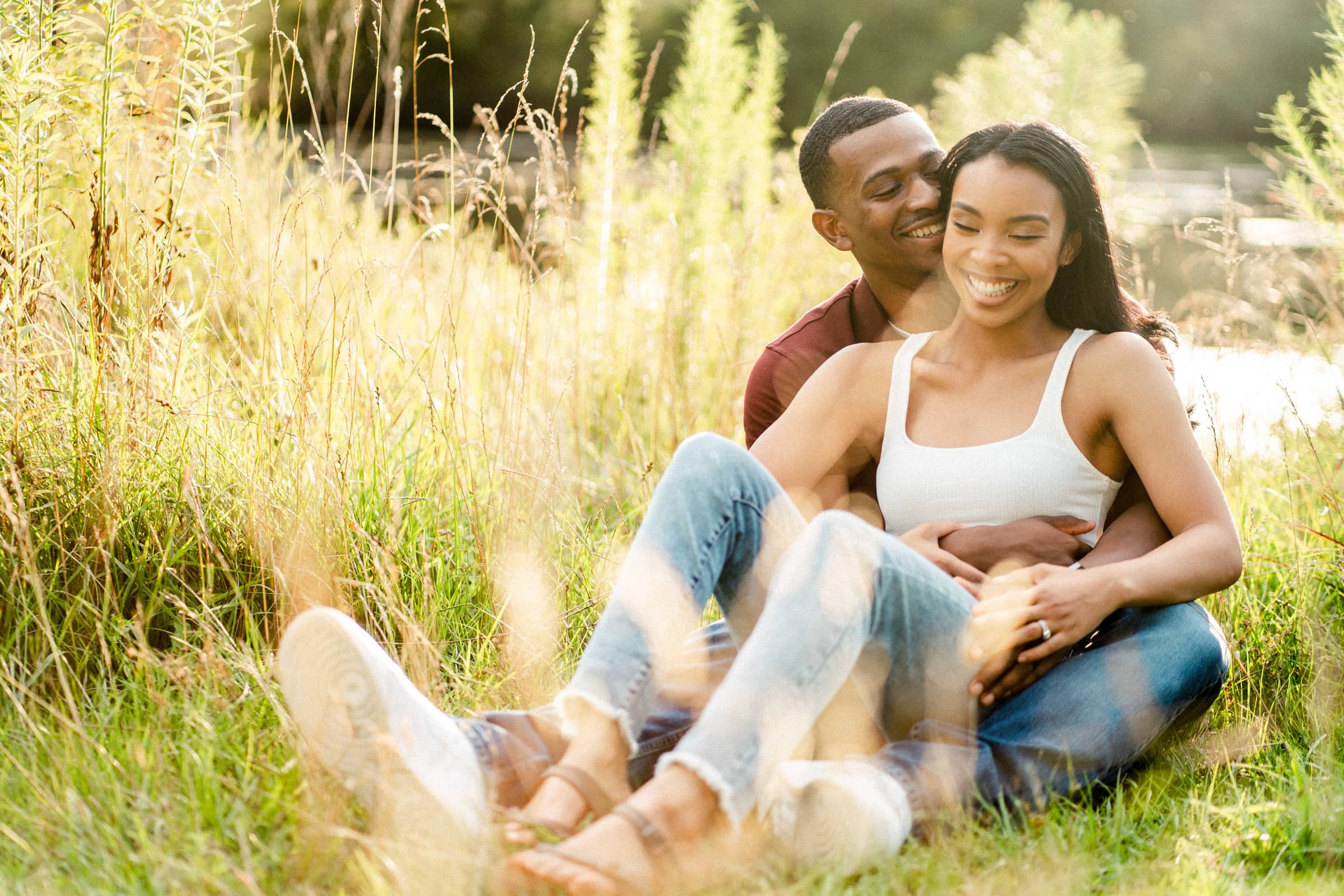 raleigh engagement photographer - kayla & deandre A couple is sitting together in a sunlit grassy field. They are smiling and embracing, with the woman sitting in front of the man. The scene is peaceful, with soft sunlight filtering through the grass.