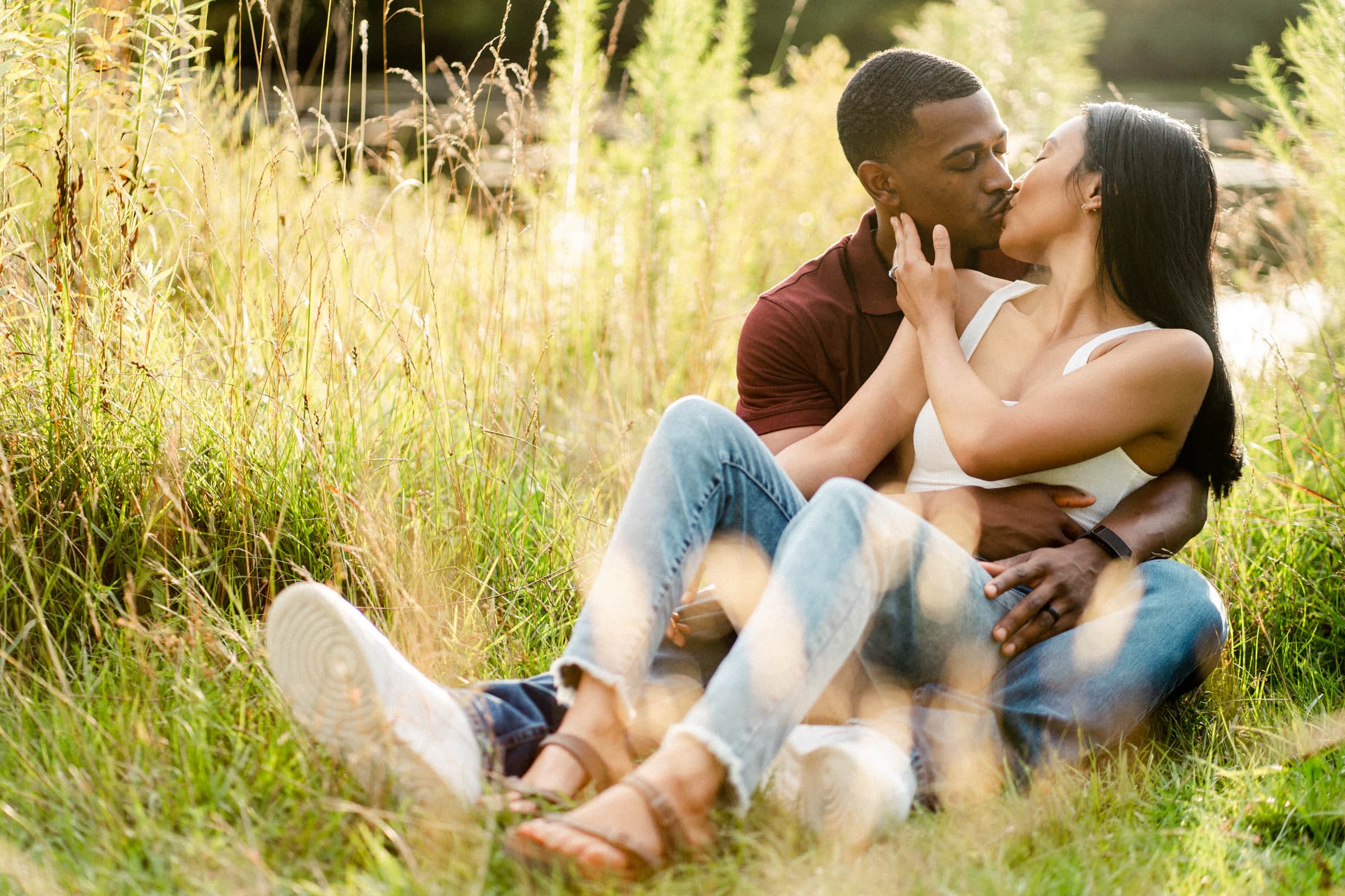 raleigh engagement photographer - kayla & deandre A couple sitting in a grassy field, kissing. The man wears a maroon shirt and the woman a white tank top with jeans. They are surrounded by tall grass under warm sunlight.