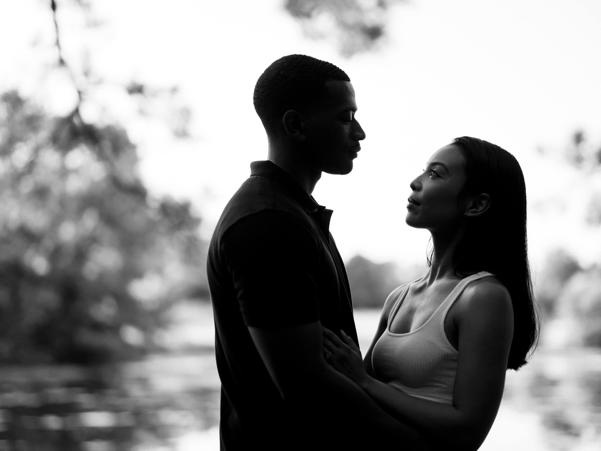 raleigh engagement photographer - kayla & deandre A black and white image of a couple standing close, gazing into each others eyes. The background is blurred, with trees and water, creating a serene atmosphere. The woman has long hair and the man has short hair.