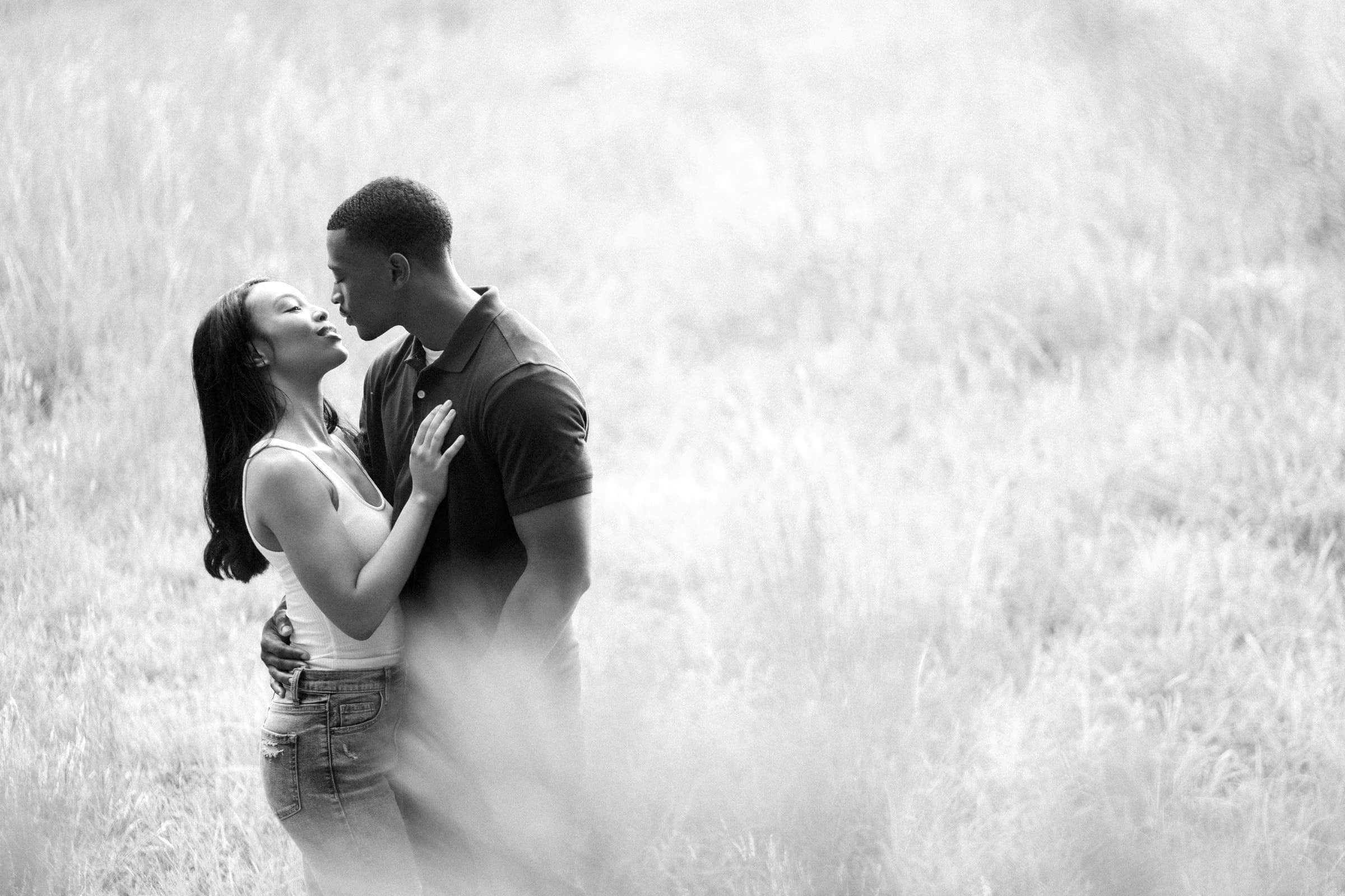 raleigh engagement photographer - kayla & deandre A black and white photograph of a couple standing in a grassy field, embracing and gazing into each others eyes. The woman has long hair and is wearing a tank top, while the man wears a short-sleeved shirt. The scene evokes a sense of romance and tranquility.