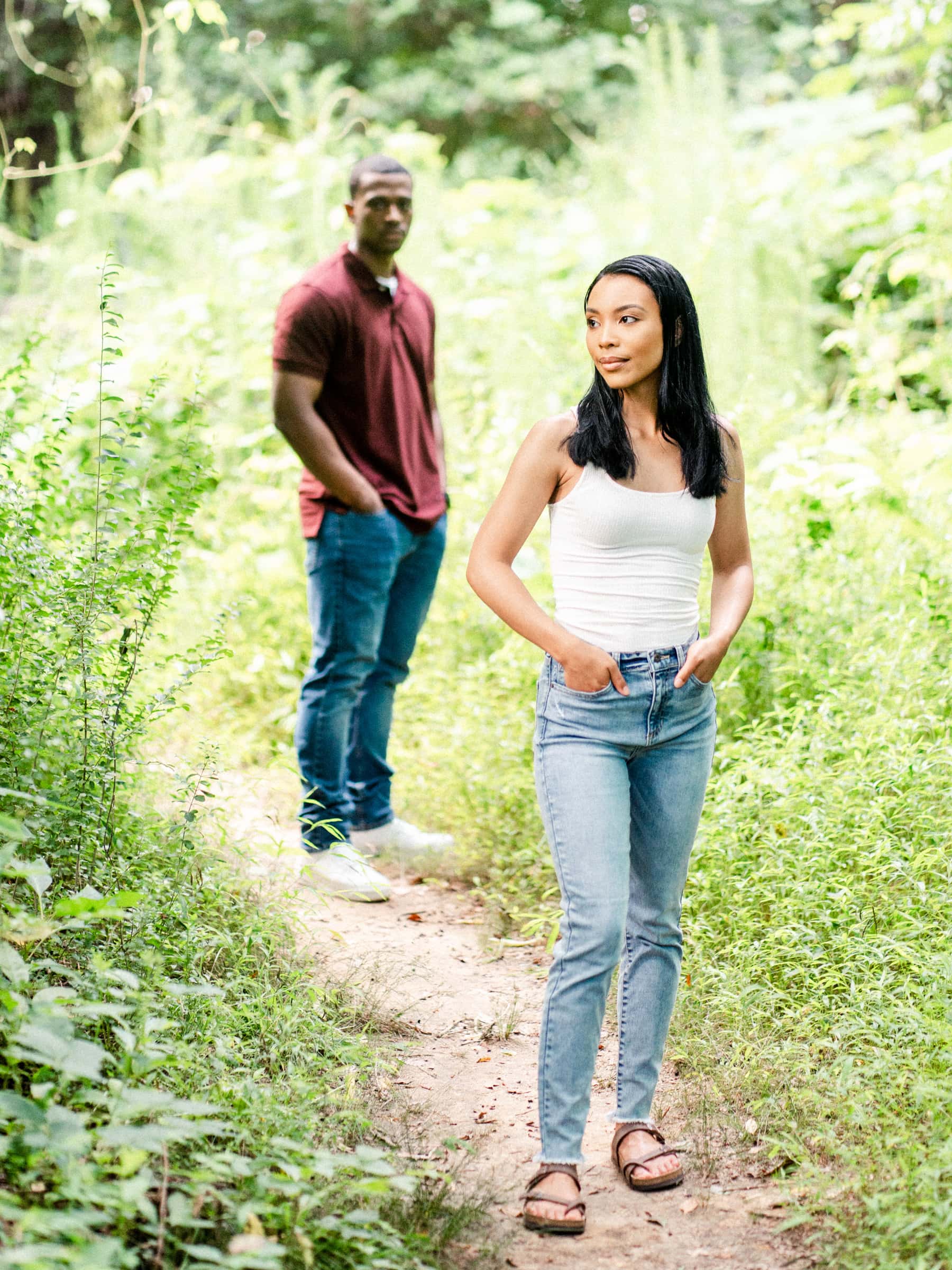 raleigh engagement photographer - kayla & deandre A woman in a white tank top and jeans stands on a forest path with her hands in her pockets, looking to the side. A man in a maroon shirt and jeans stands in the background among green foliage.