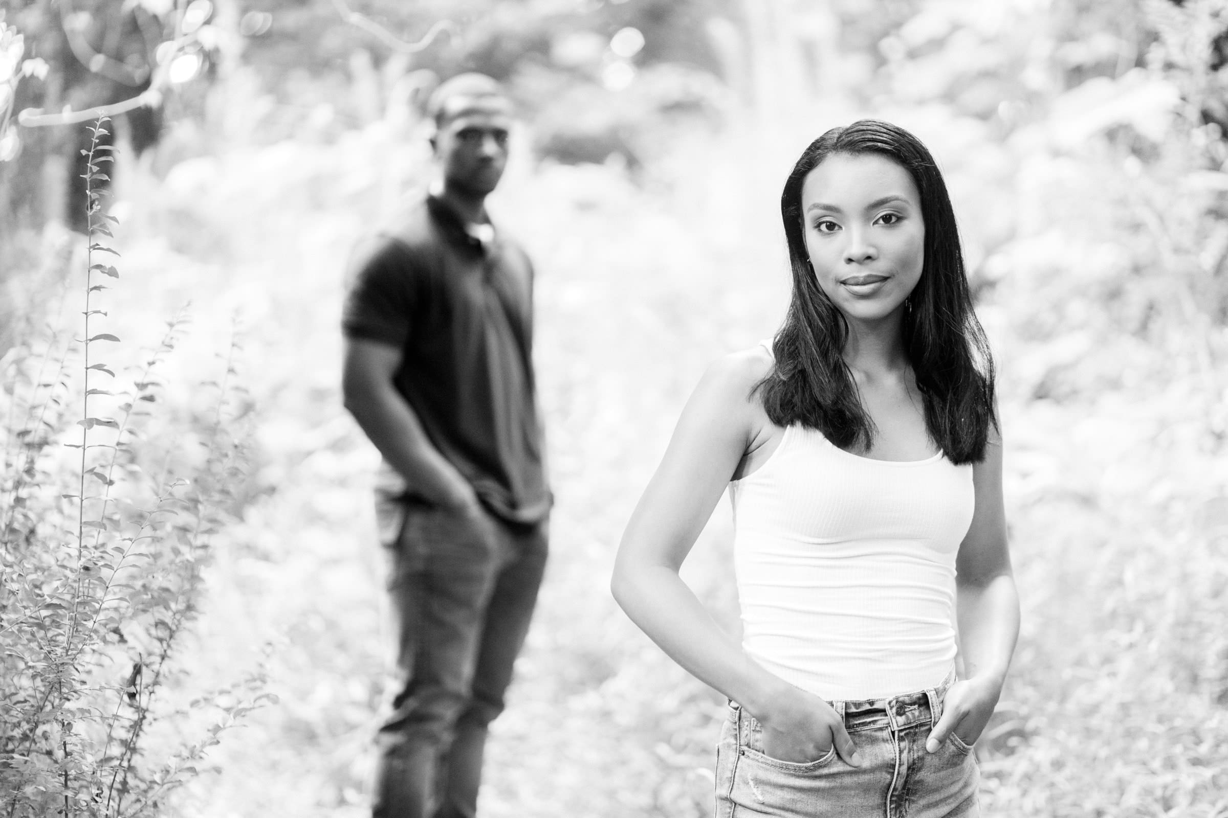 raleigh engagement photographer - kayla & deandre A black and white image of a woman in a white tank top and jeans standing in the foreground of a wooded path. She has her hands in her pockets and looks at the camera. A man stands out of focus in the background, wearing a dark shirt and pants.