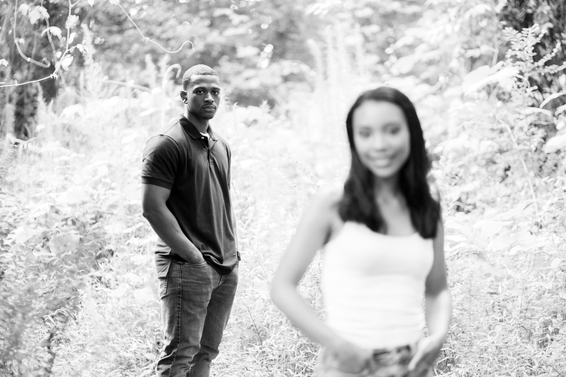 raleigh engagement photographer - kayla & deandre A black and white photo of a man in the background standing with hands in pockets, wearing a dark shirt and jeans, and a woman in the foreground out of focus, wearing a white tank top, against a lush, natural backdrop.