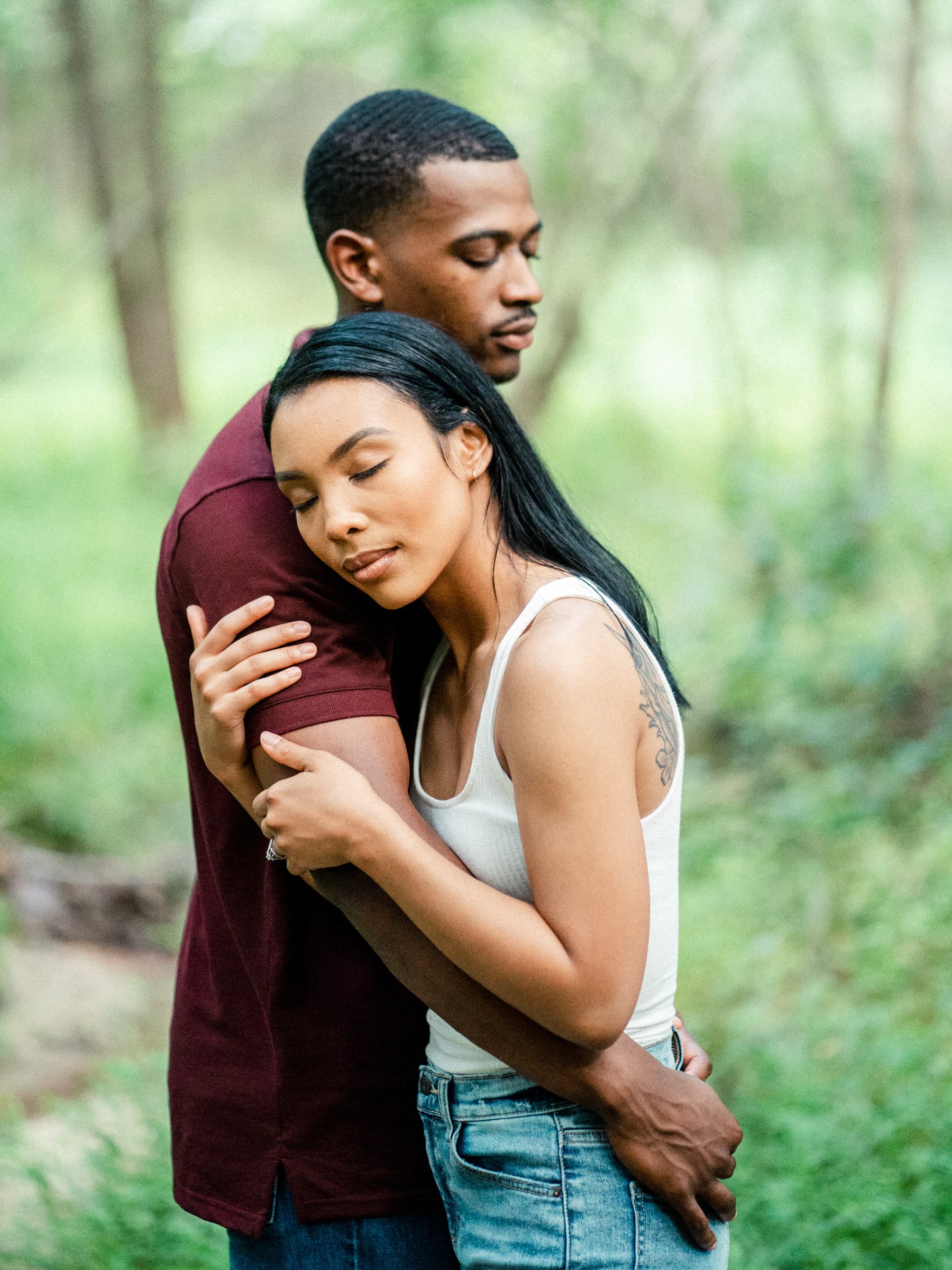 raleigh engagement photographer - kayla & deandre A couple embraces peacefully in a lush, green forest. The woman, wearing a white tank top and jeans, rests her head on the mans shoulder. The man, in a burgundy shirt, gently holds her in a calm and tranquil setting.