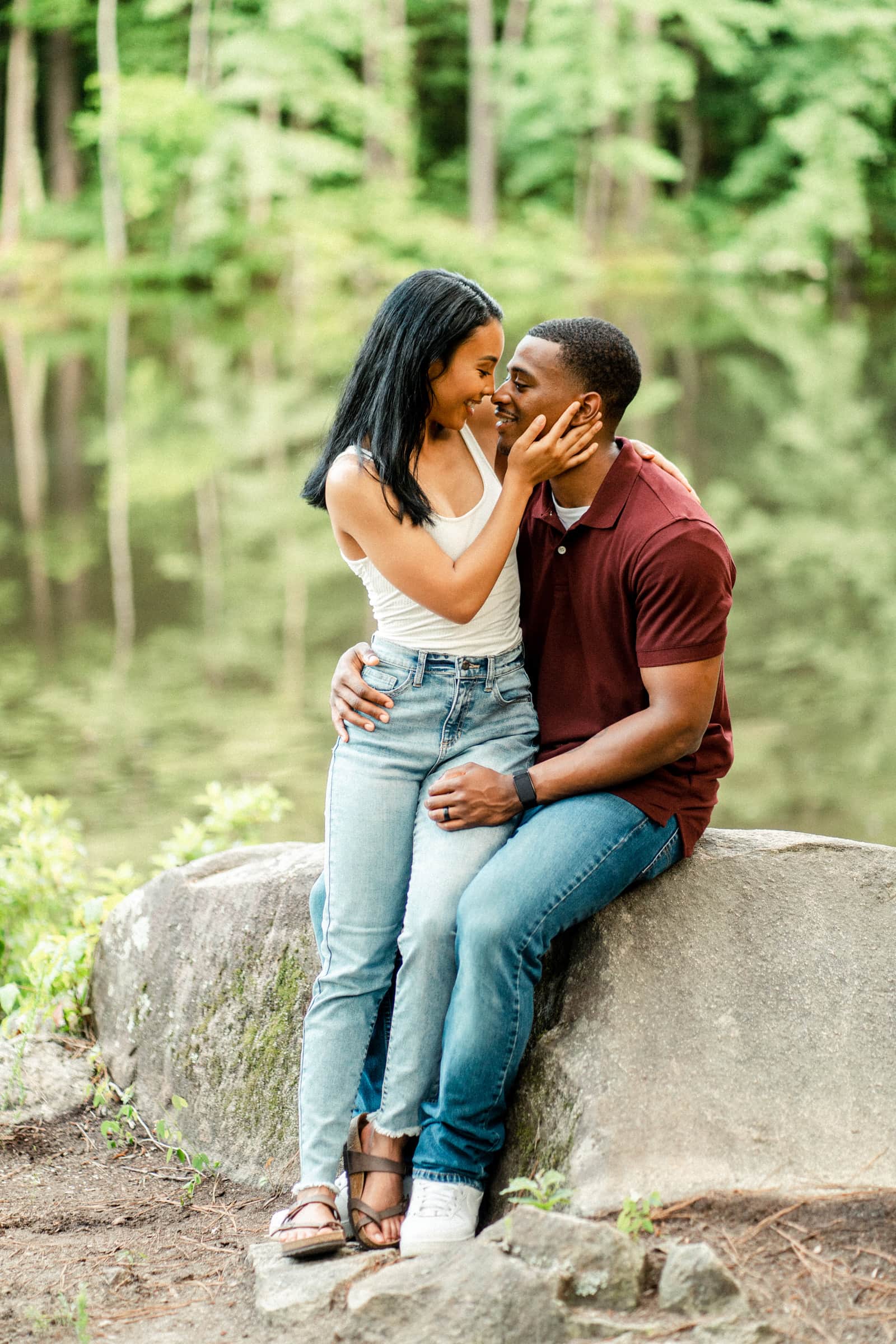 raleigh engagement photographer - kayla & deandre A couple sits on a rock by a tranquil pond in a forest. The woman, wearing a white tank top and jeans, lovingly holds the mans face. The man, in a maroon shirt and jeans, embraces her waist. The background is lush with greenery and calm water.