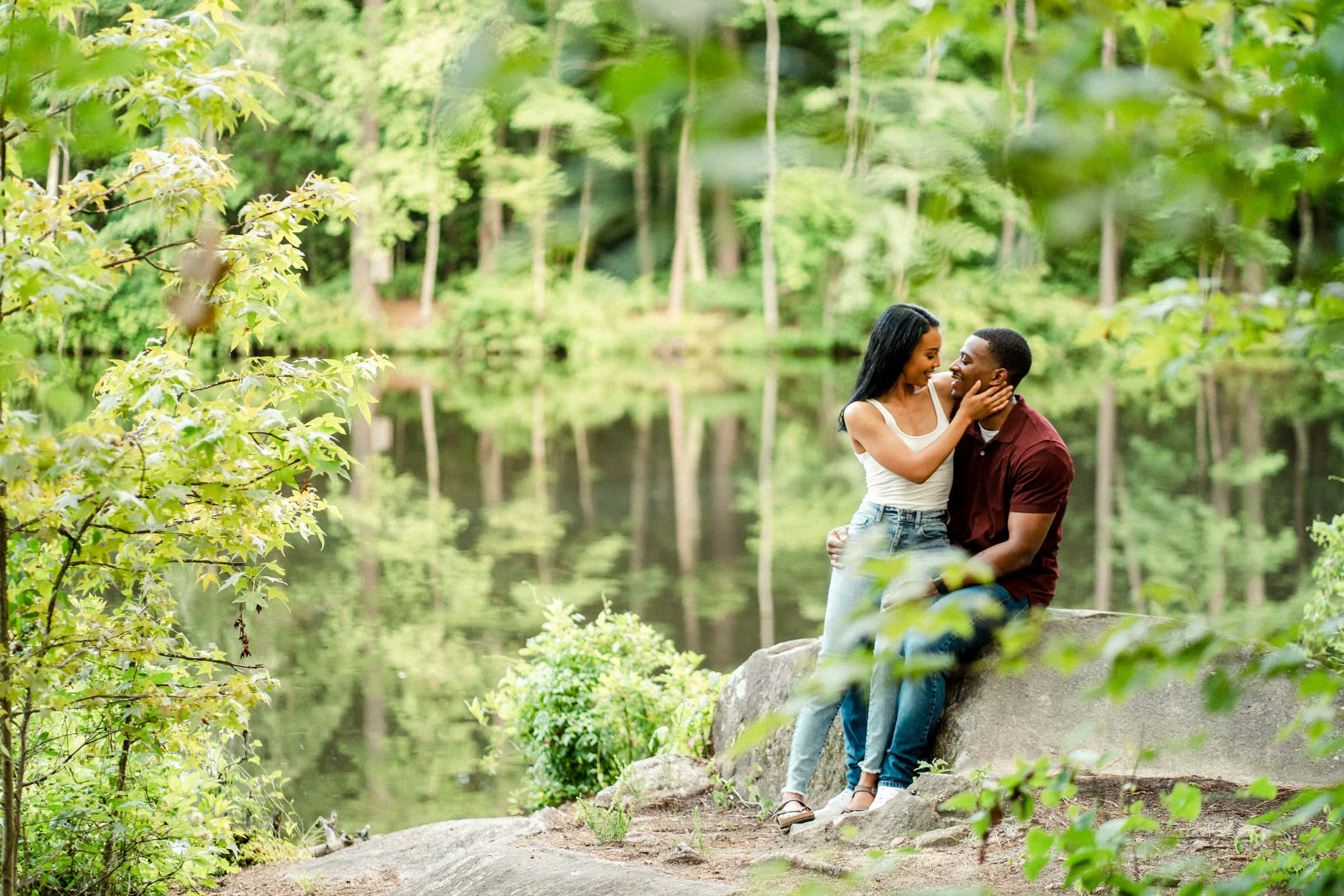 raleigh engagement photographer - kayla & deandre A couple sits on a large rock by a serene, reflective lake surrounded by lush greenery. The woman tenderly touches the mans face as they gaze at each other affectionately, creating a romantic scene in nature.