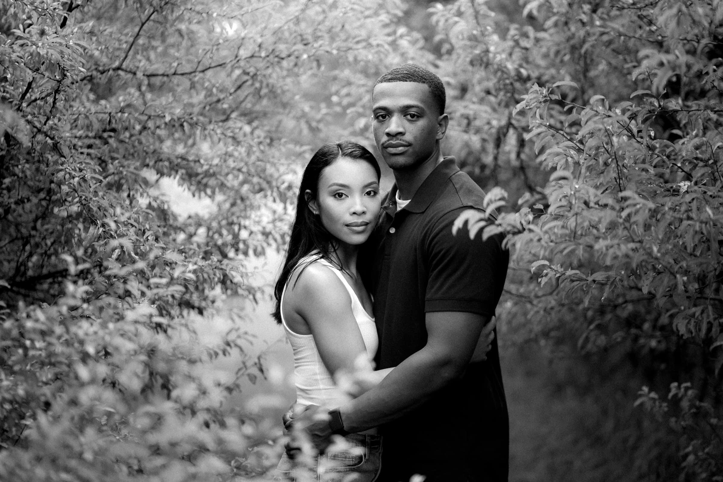 raleigh engagement photographer - kayla & deandre A black and white photo of a couple standing closely and embracing in a wooded area. They are surrounded by dense foliage, and the man is wearing a dark shirt while the woman is in a light tank top. They gaze directly at the camera.