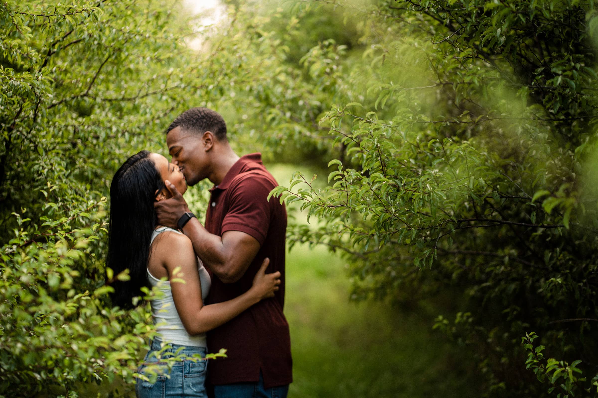 raleigh engagement photographer - kayla & deandre A couple embraces and kisses in a lush, green pathway surrounded by dense foliage. The man is wearing a maroon shirt and the woman is in a white tank top and denim shorts. Sunlight filters through the leaves, creating a serene atmosphere.